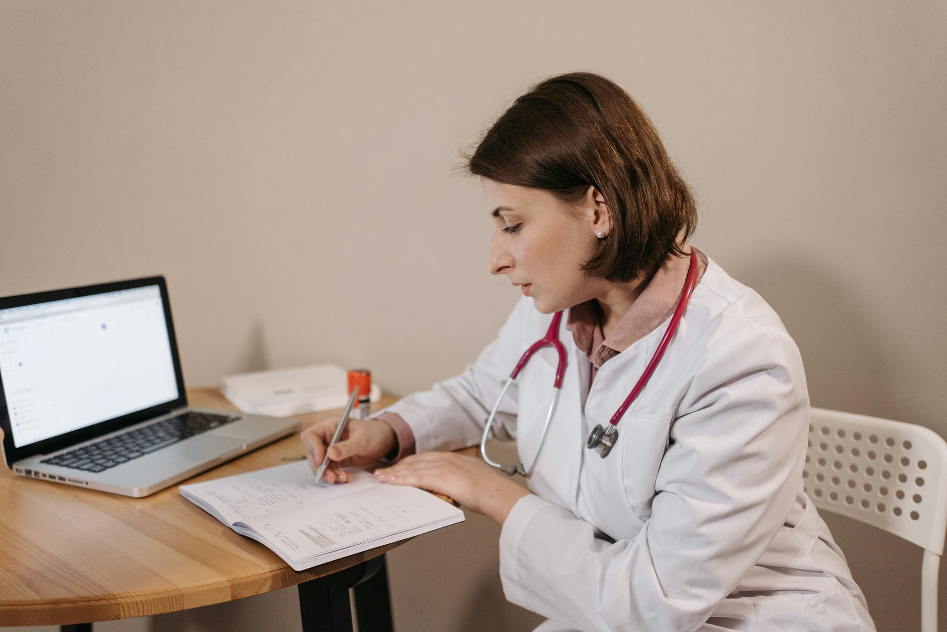 Doctor in white coat, stethoscope, writing at a desk with laptop and notebook.