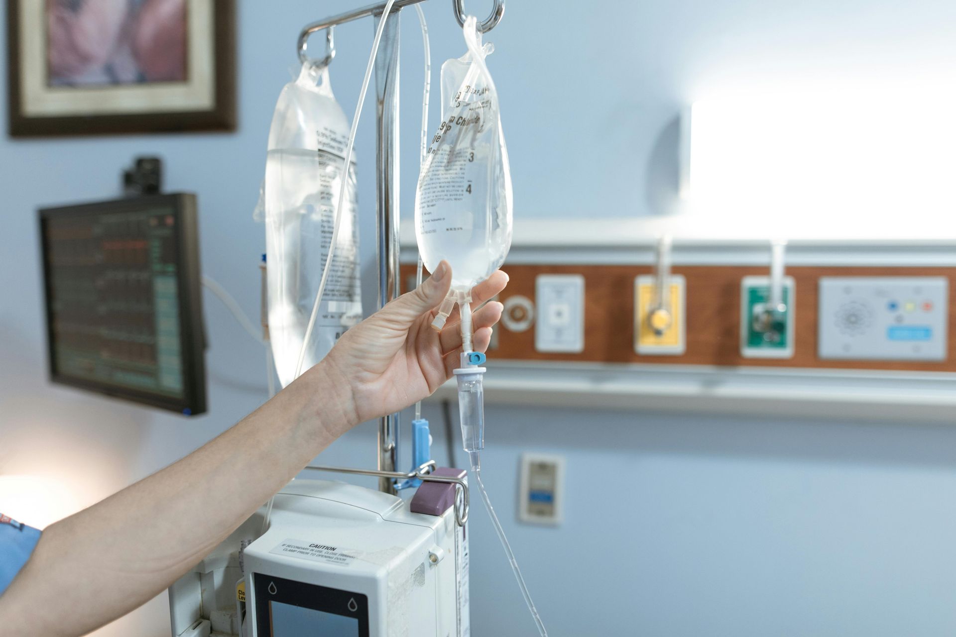 Hand adjusting IV bag in a hospital room. IV pole and medical equipment are in the frame.
