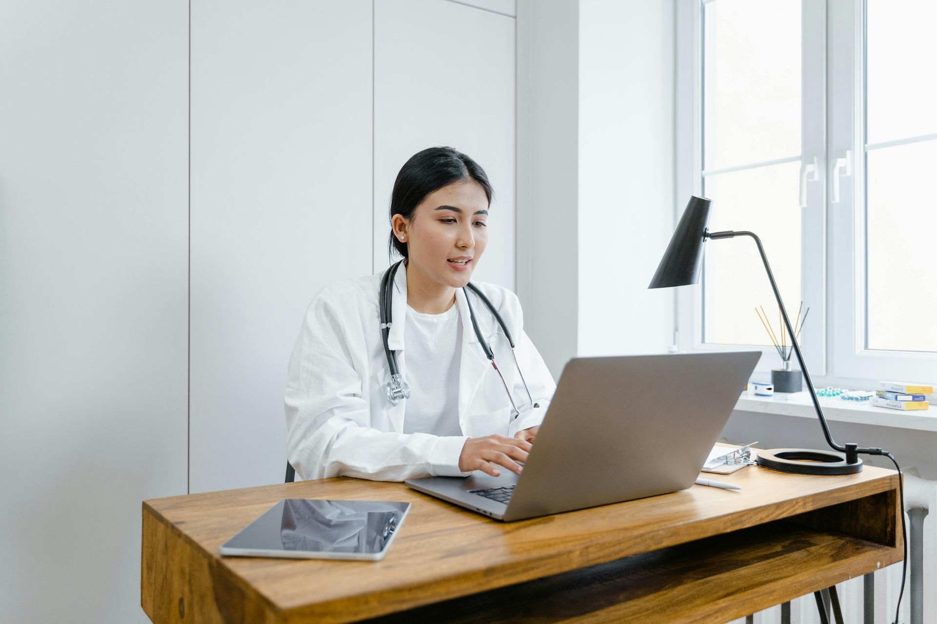 Doctor in white coat with stethoscope, typing on laptop at a desk with tablet near window.