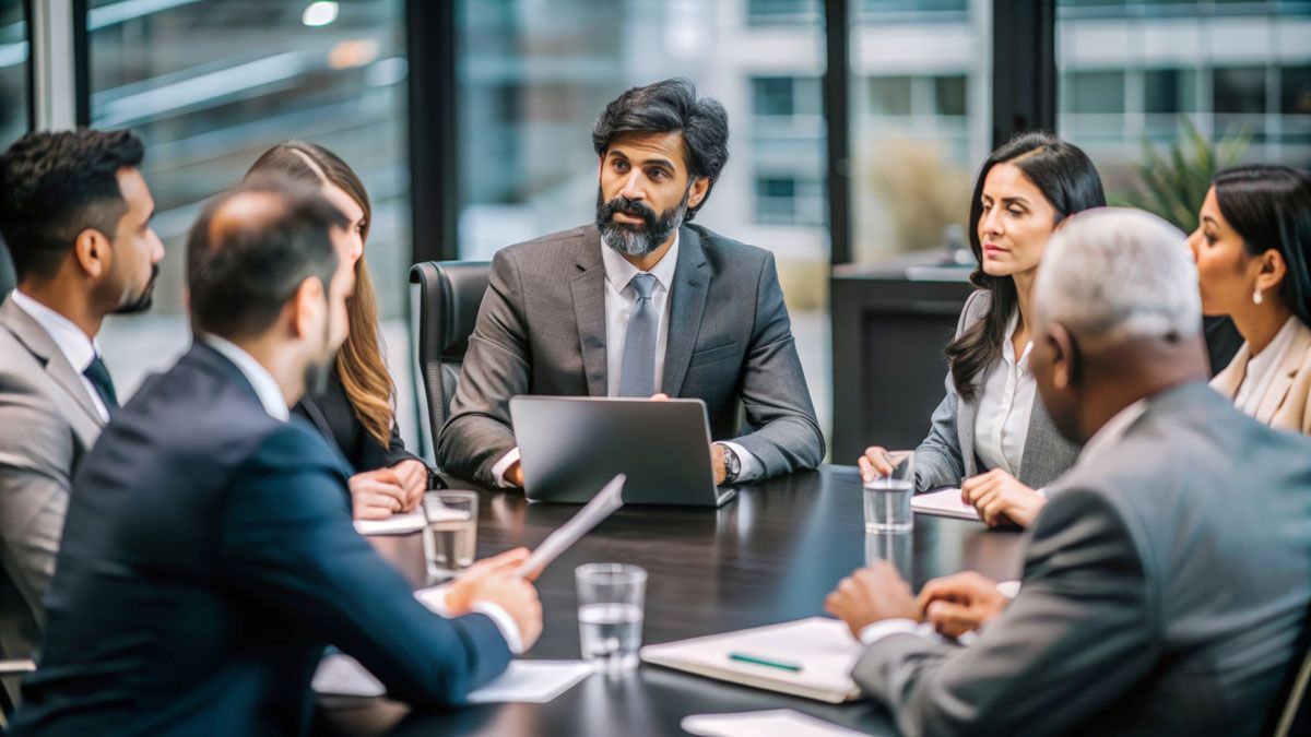 Business meeting: diverse group around a conference table, discussing, laptop in center.