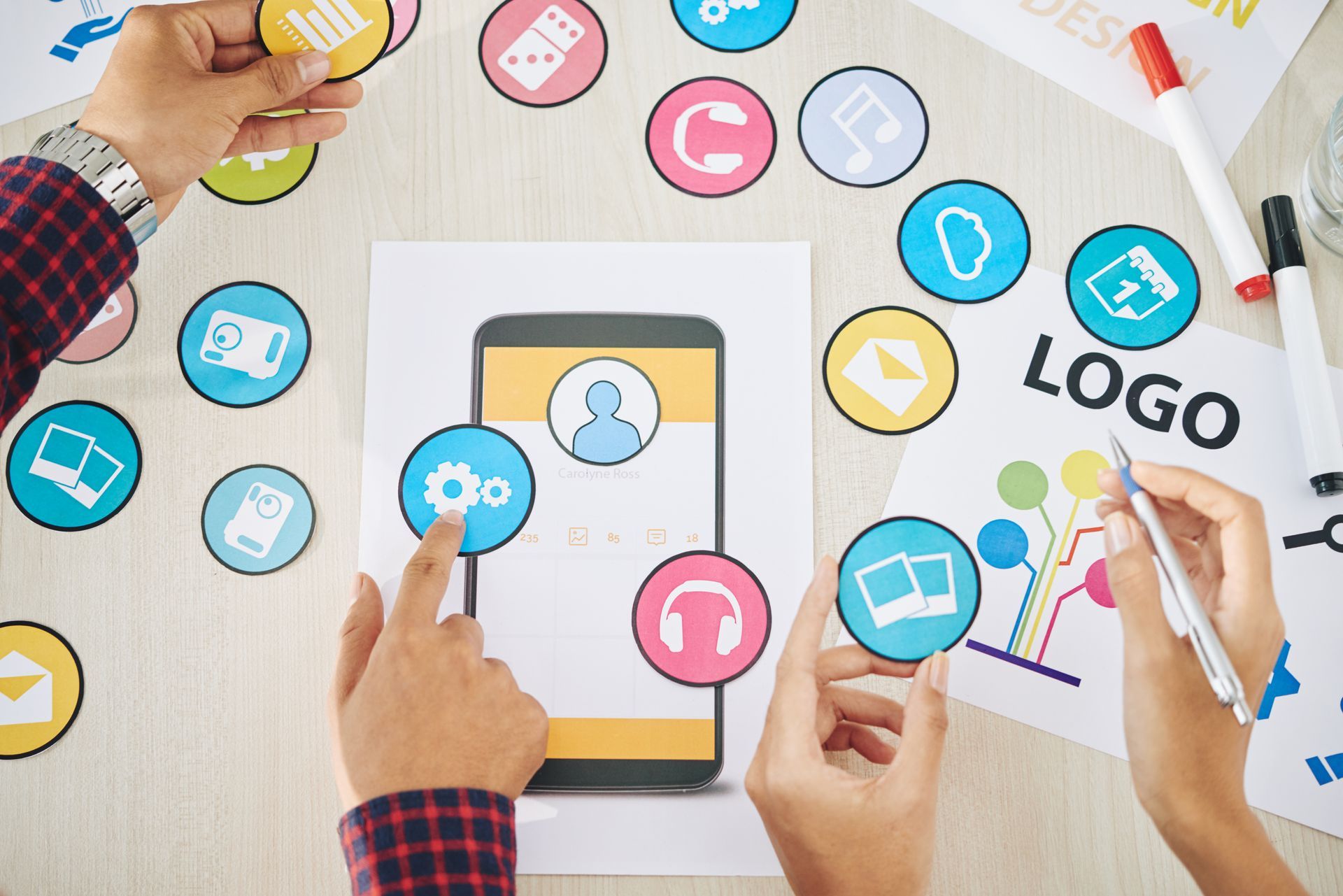 Hands designing app interface on smartphone, colorful icons and logo on desk.