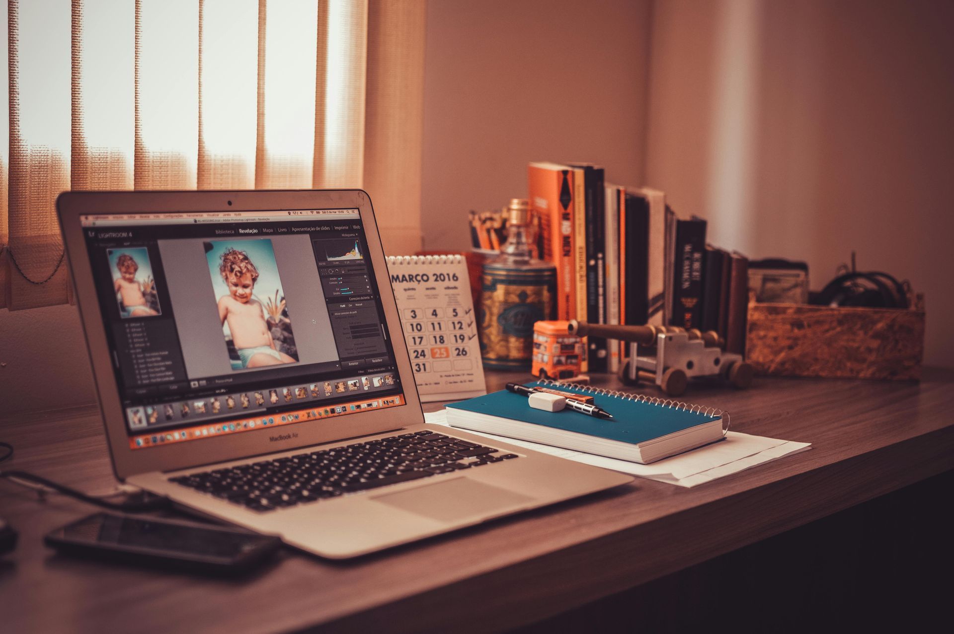 Laptop on a desk with a photo editing program open. A child's photo is visible. Books, calendar, notepad.