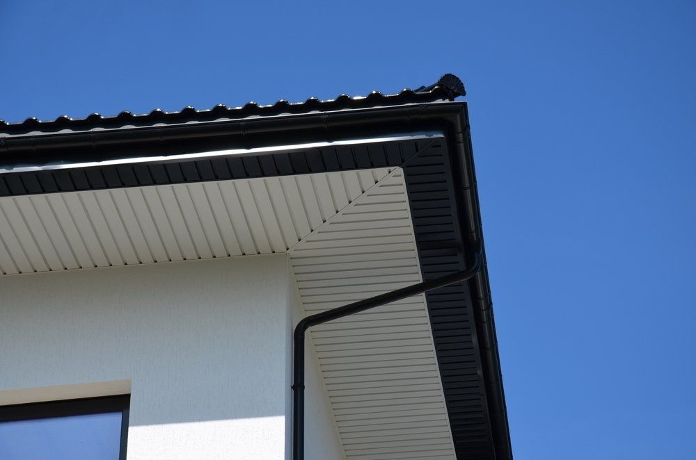 Coin d'une maison avec toit, gouttières et moulures noirs. Revêtement clair sur fond de ciel bleu.