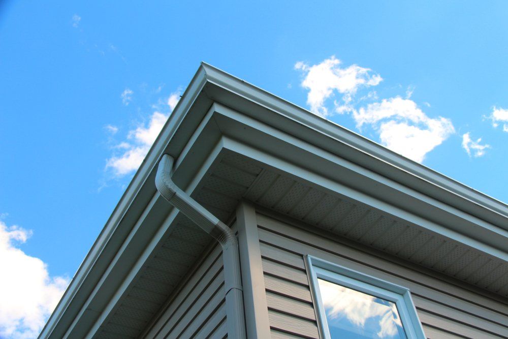 Coin d'une maison avec un revêtement marron clair, des garnitures blanches et une gouttière contre un ciel bleu avec des nuages.