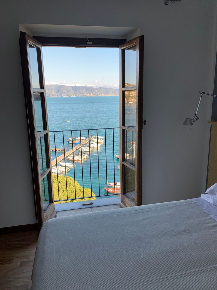 Finestra aperta con vista su un lago con un letto in primo piano. Si vedono l'acqua blu, le montagne e un piccolo porto.