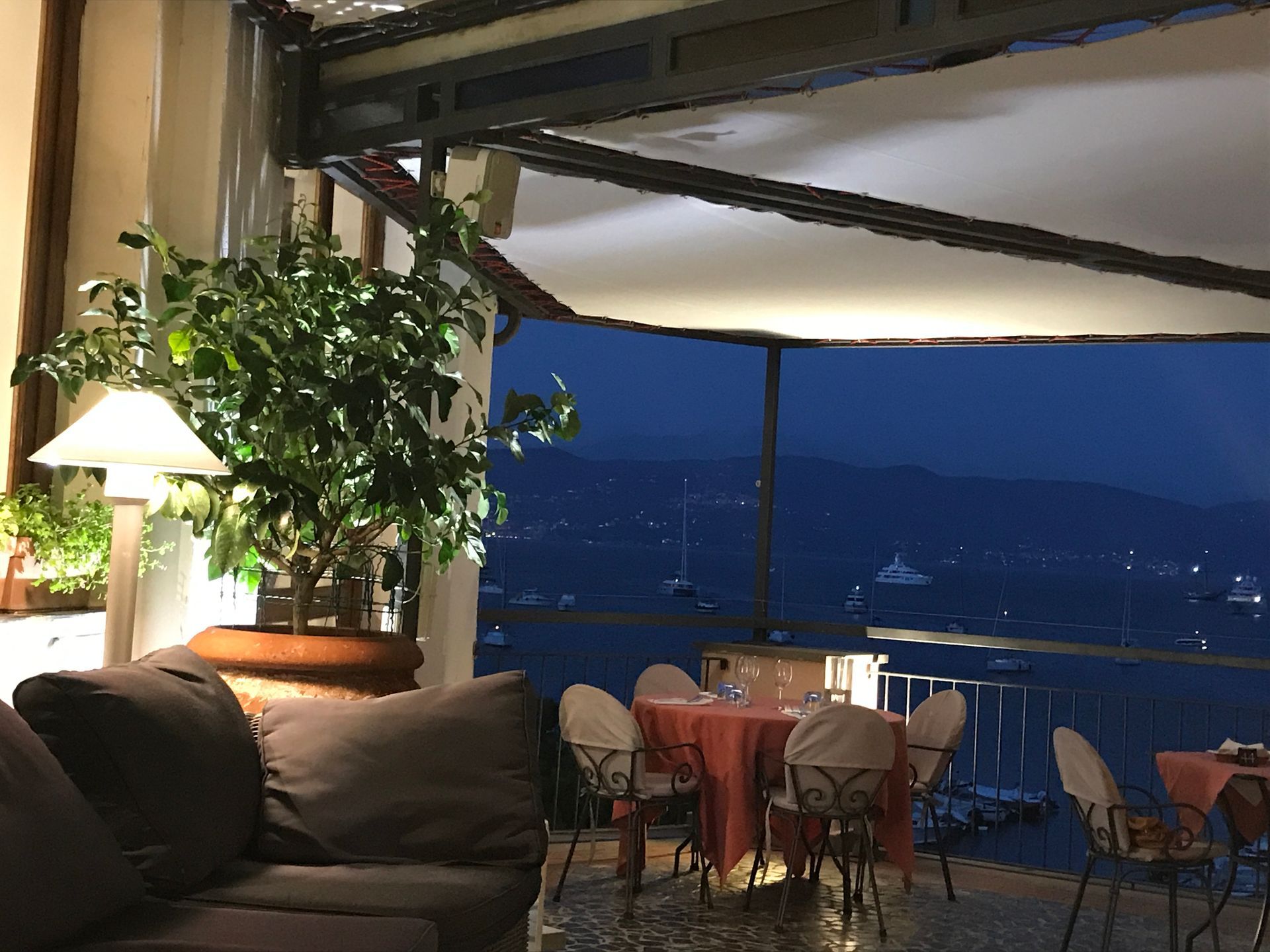Patio del ristorante con vista sul porto buio; tavolo apparecchiato con sedie, grande pianta in vaso e divano.