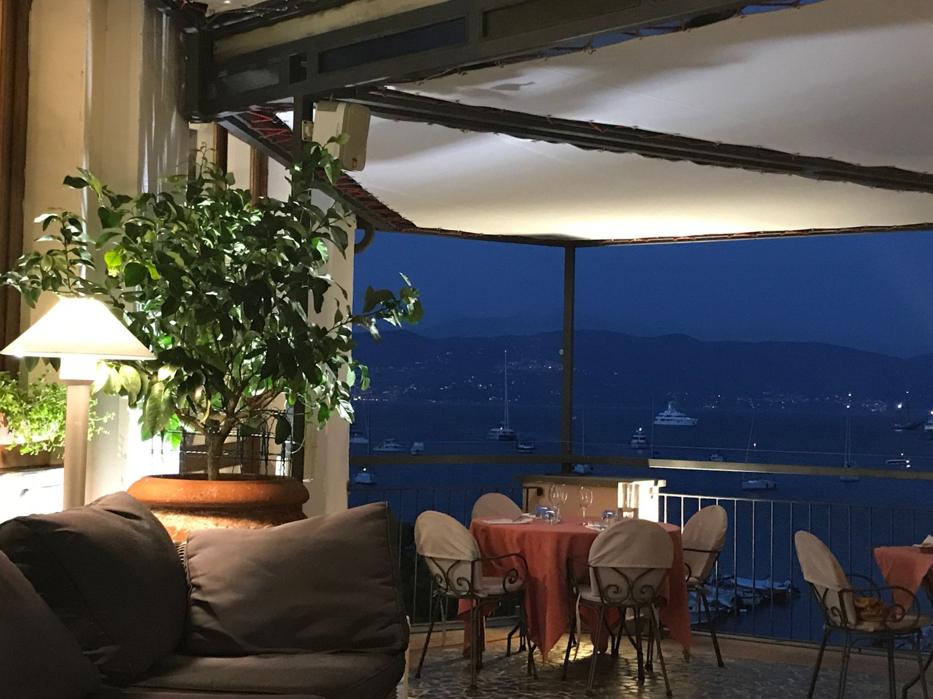 Ristorante all'aperto al tramonto, con tavoli apparecchiati, con vista sul mare scuro e luci lontane.