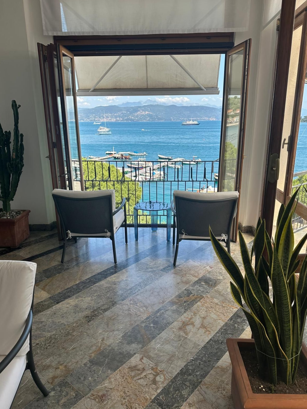Due sedie su un balcone con vista sul porto turistico e sul mare, incorniciato da porte aperte.
