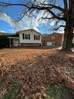 A white two-story house with a brick garage adjacent, viewed from a yard covered in fallen brown leaves under a sunny sky.