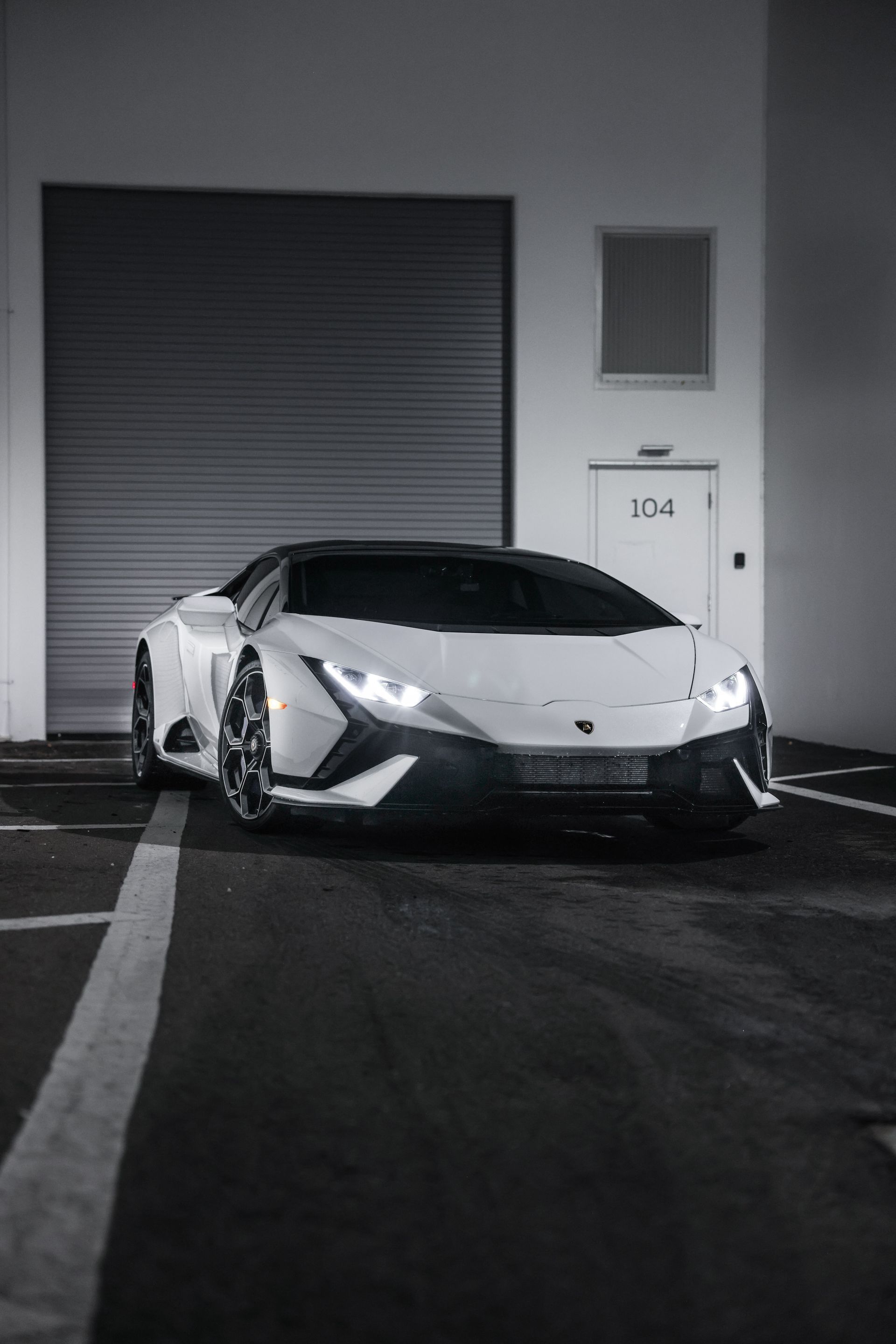 White sports car parked at night beside a closed garage door and building entrance marked 104