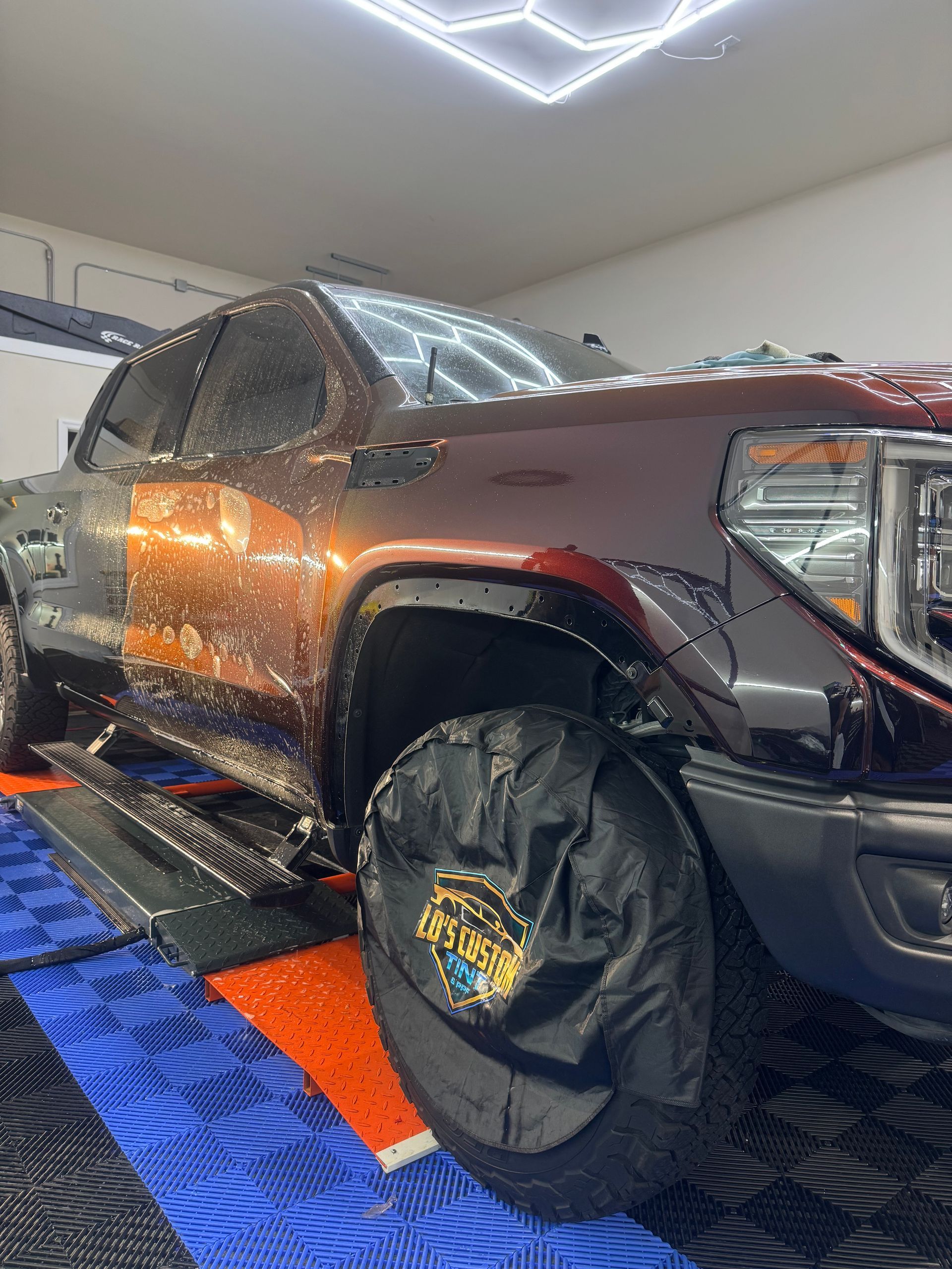 Black pickup truck indoors, covered in reflective silver wrap with a spare tire cover and patterned floor mats.