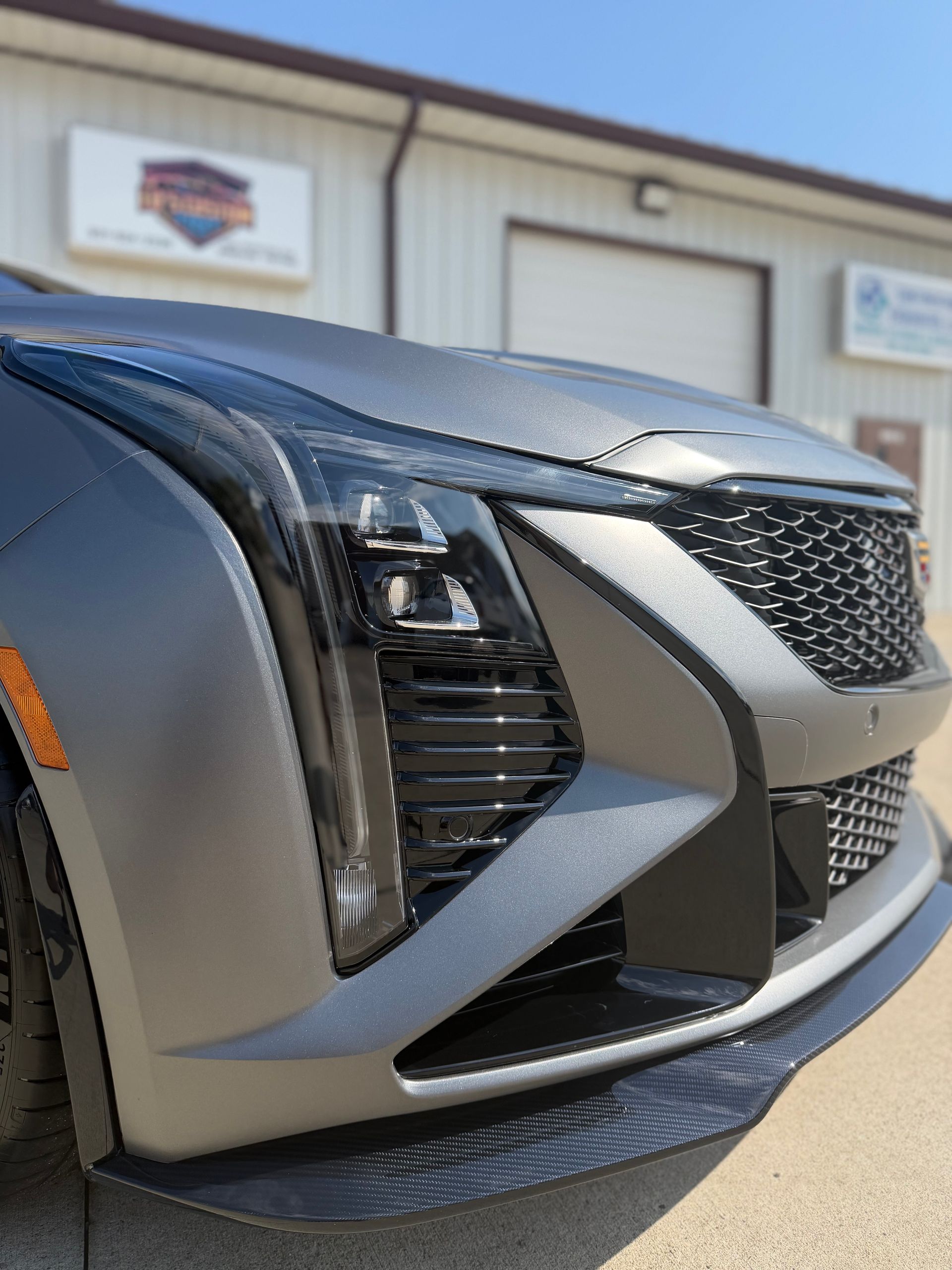 Close-up of a gray car front grille and headlight outside a building with garage doors.
