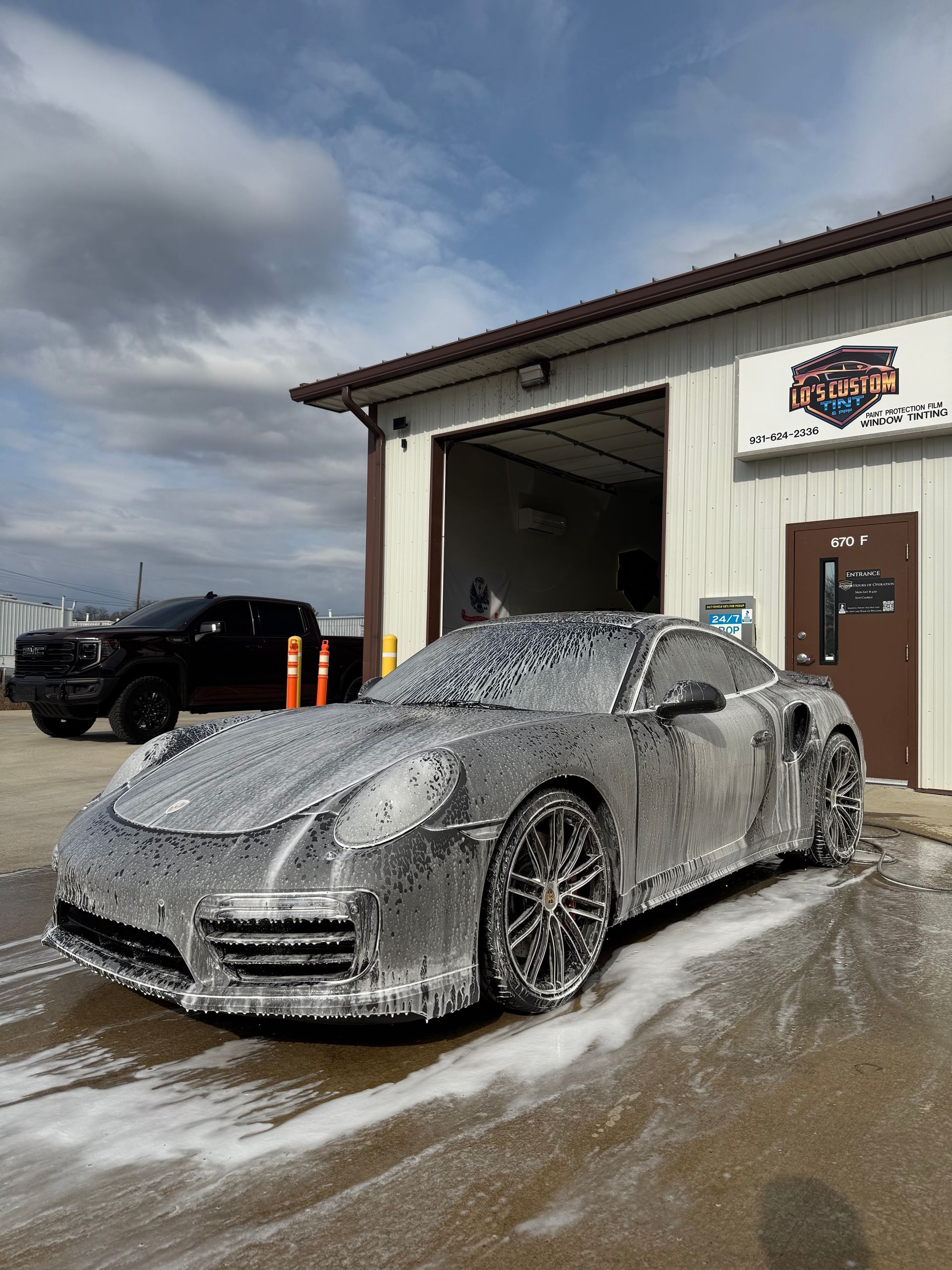 Snow-covered gray sports car outside a garage on a wet lot under cloudy skies