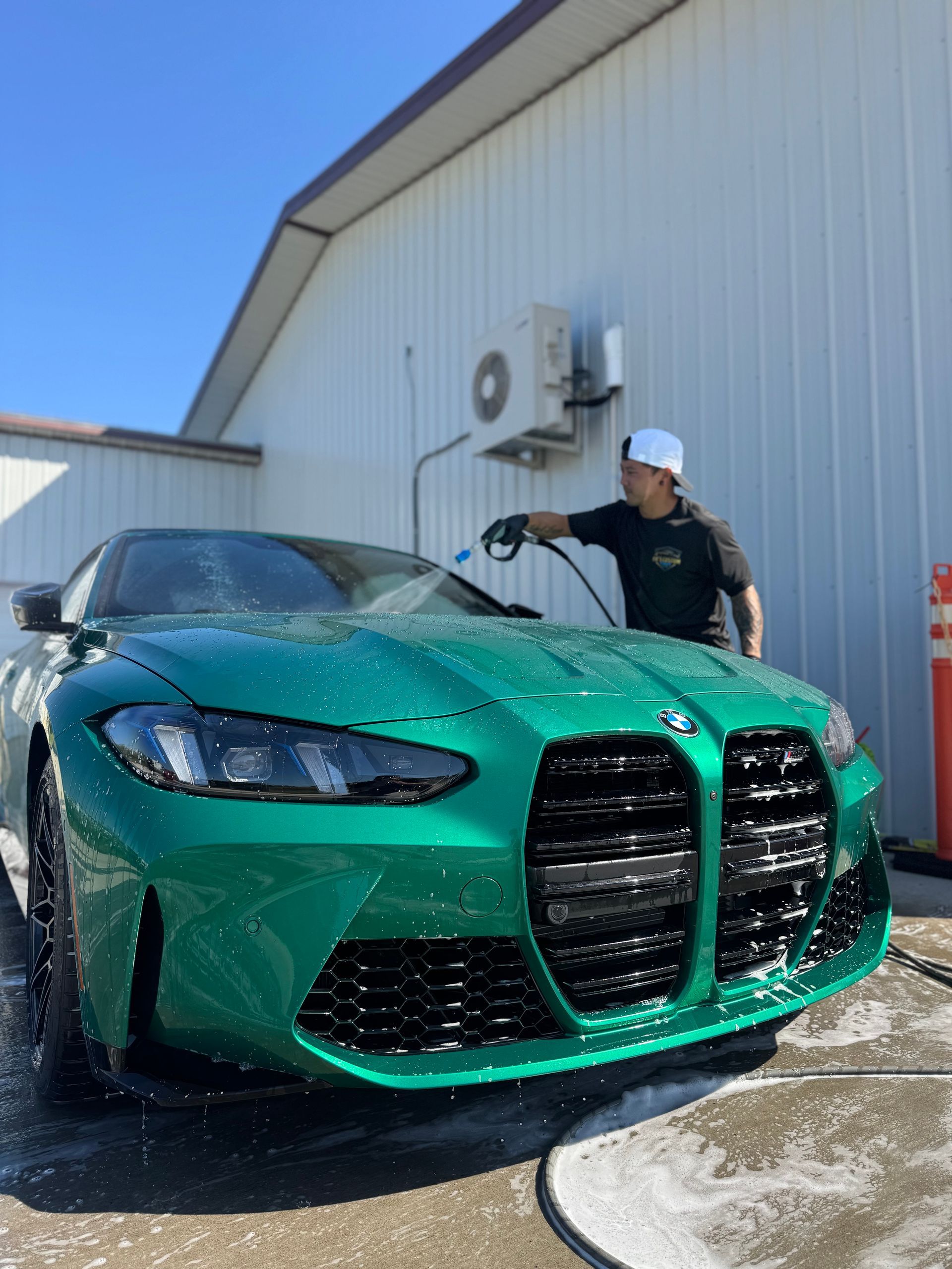 Green BMW being washed by a person outside a white building on a sunny day