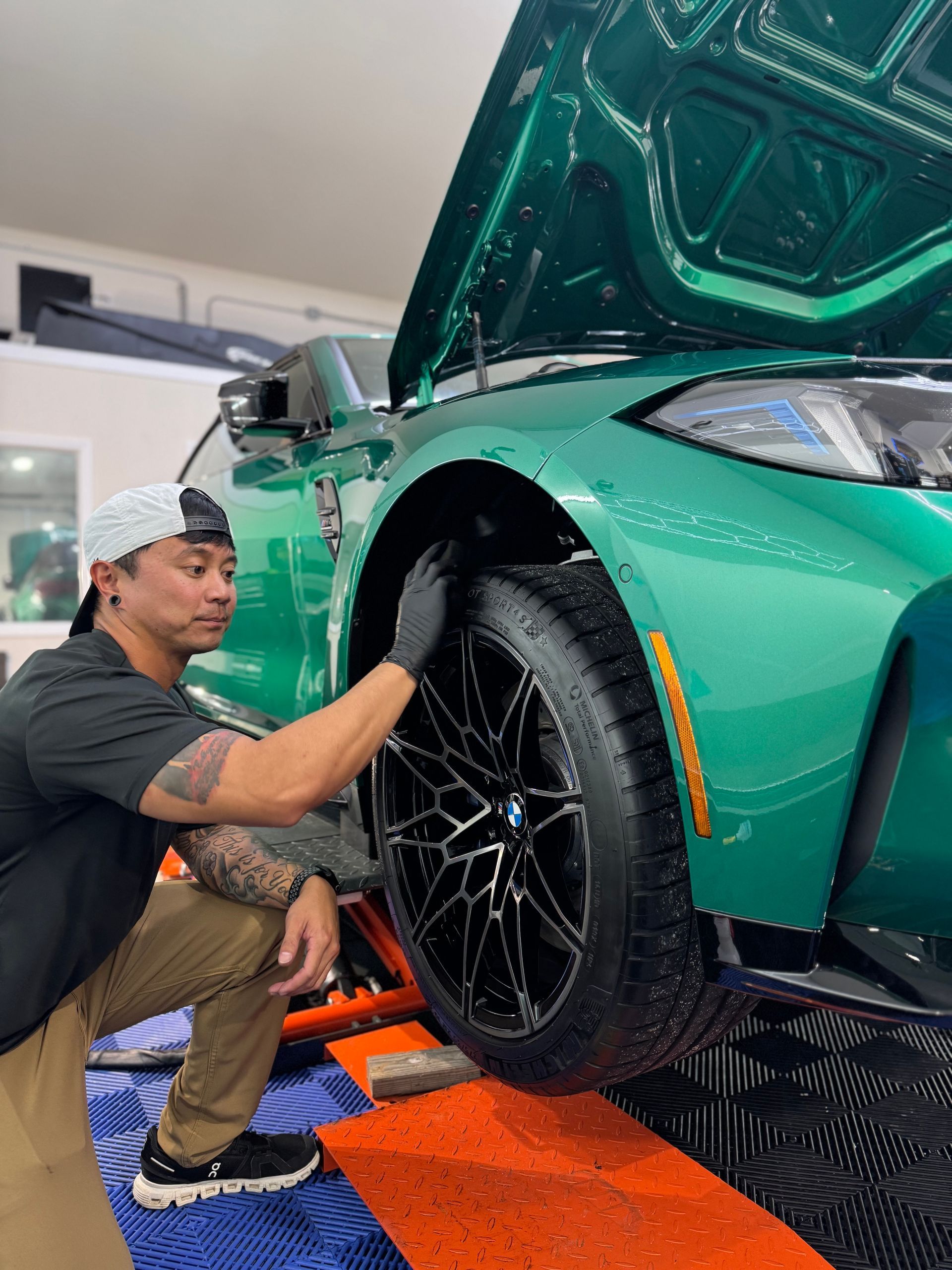 Mechanic inspecting a teal sports car’s front wheel in a garage, hood open.