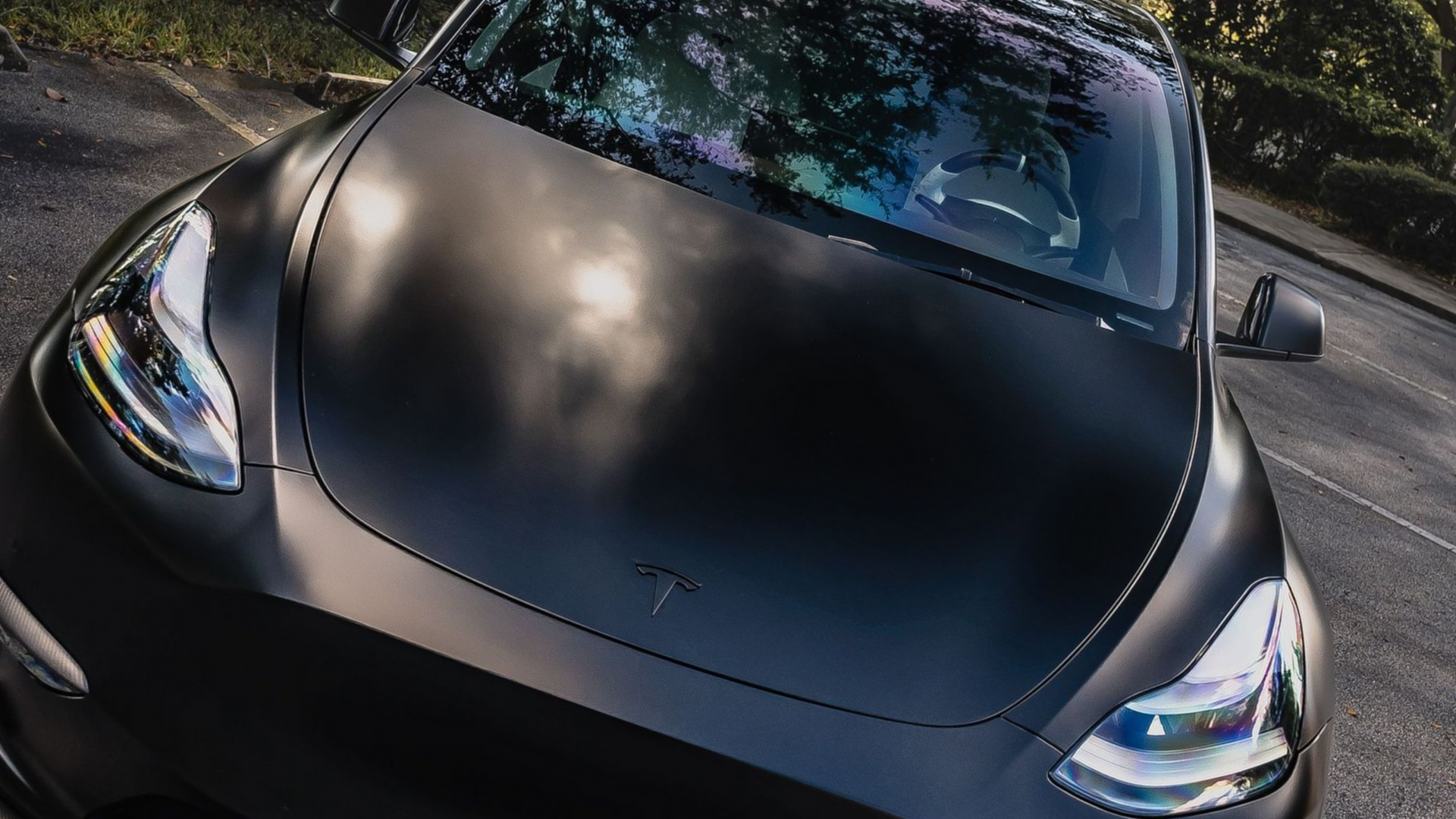 Black Tesla car hood with headlights visible, parked outdoors in sunlight.