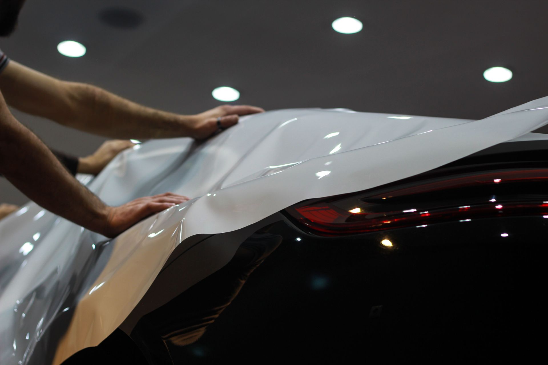 Hands applying white protective film to a black car under showroom lights