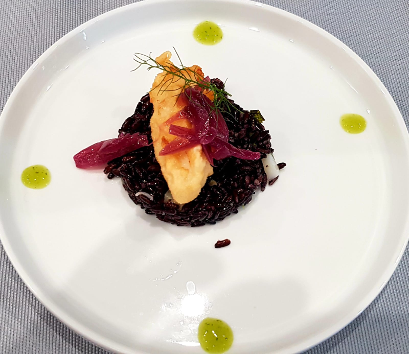 Piatto di pesce gourmet con risotto scuro, guarnizione di barbabietola e gocce di salsa verde su un piatto bianco.