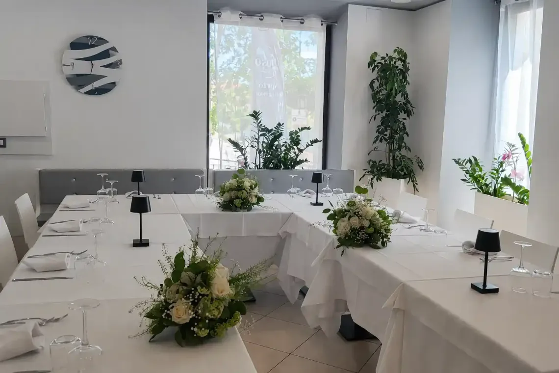 Sala da banchetto luminosa e bianca con lunghi tavoli, centrotavola floreali e grandi finestre.