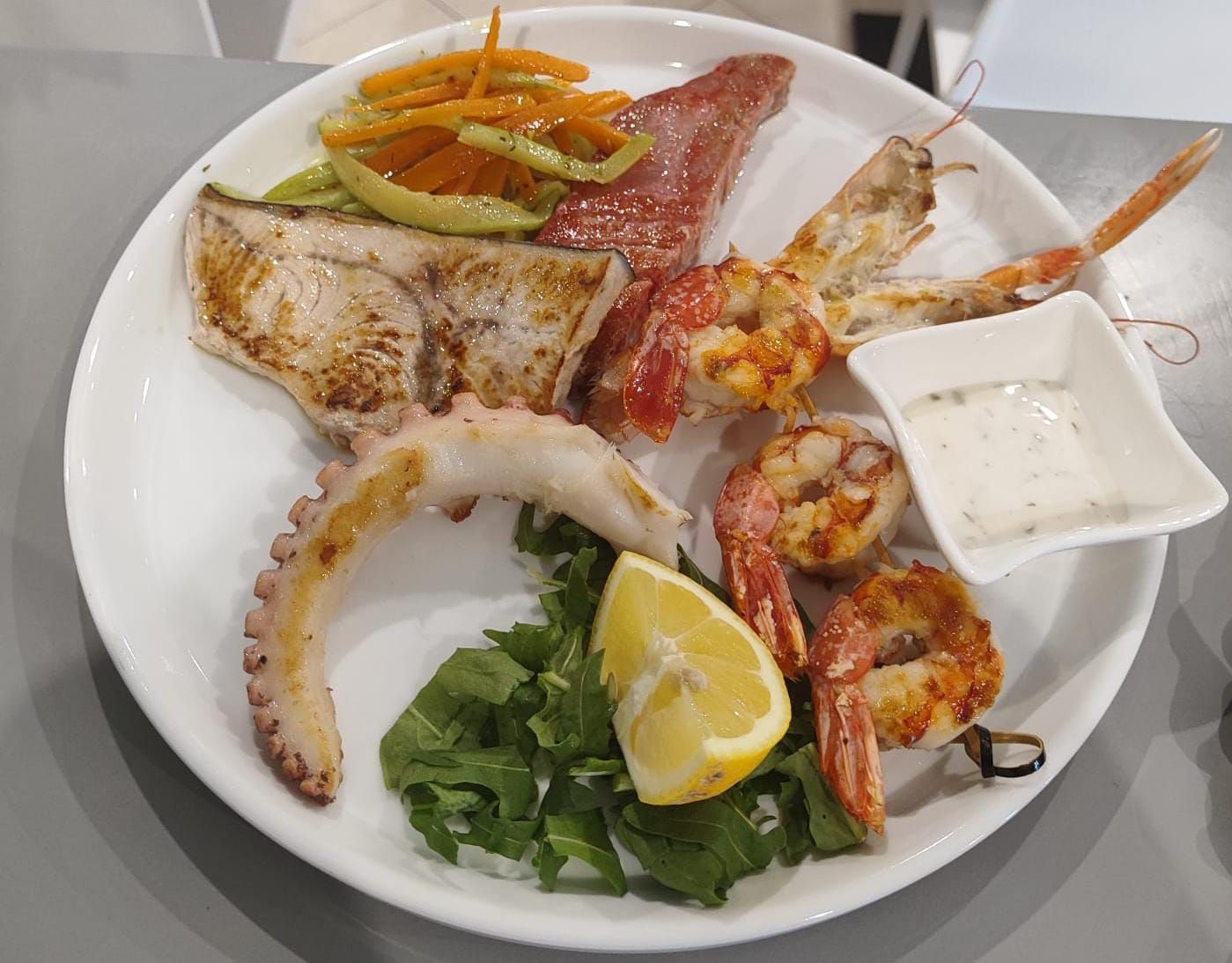 Piatto di frutti di mare con pesce grigliato, gamberi, polpo, limone, erbe aromatiche e salsa per intingere su un piatto bianco.