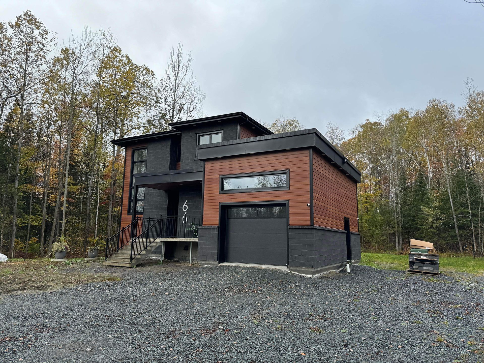 Une maison moderne avec une porte de garage noire est entourée d'arbres et de gravier.