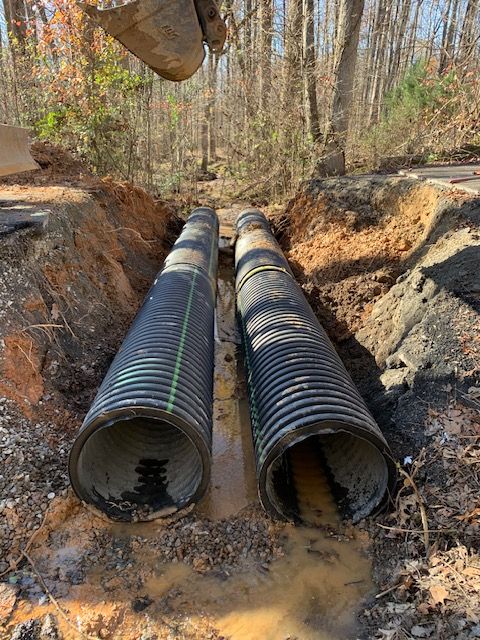 Two large black corrugated drainage pipes laid in a muddy trench in a wooded area