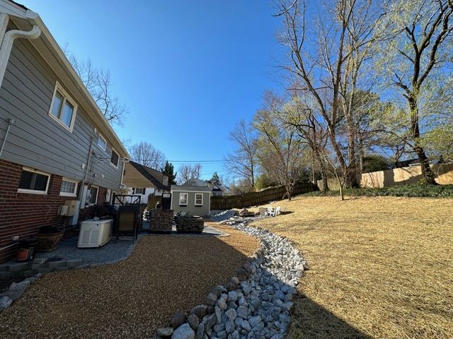 Backyard with a stone-lined curved path beside a house, lawn, trees, and a clear blue sky.