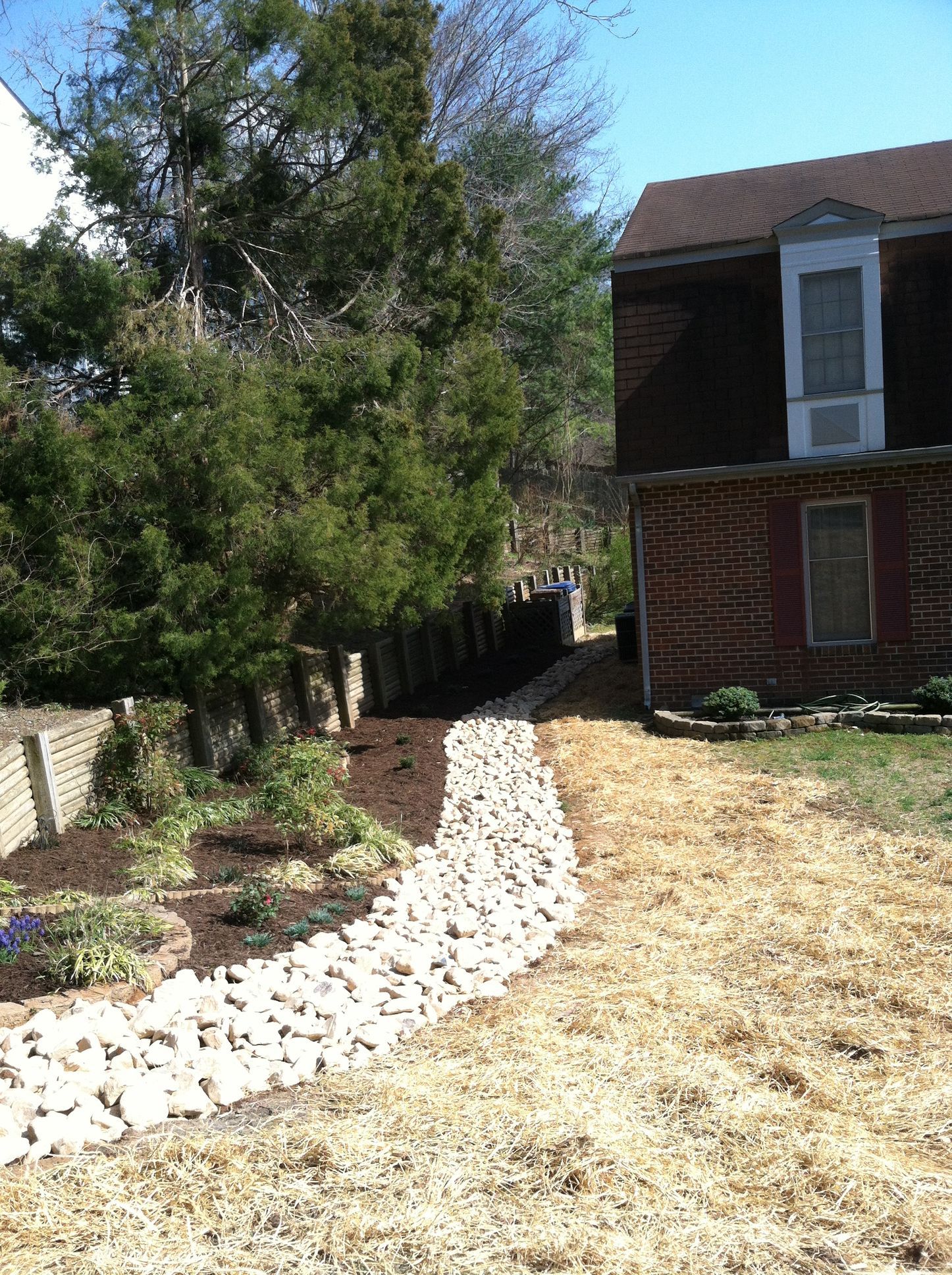 Curved white stone path beside a tree and garden leading to a brick house with a front porch and dry lawn