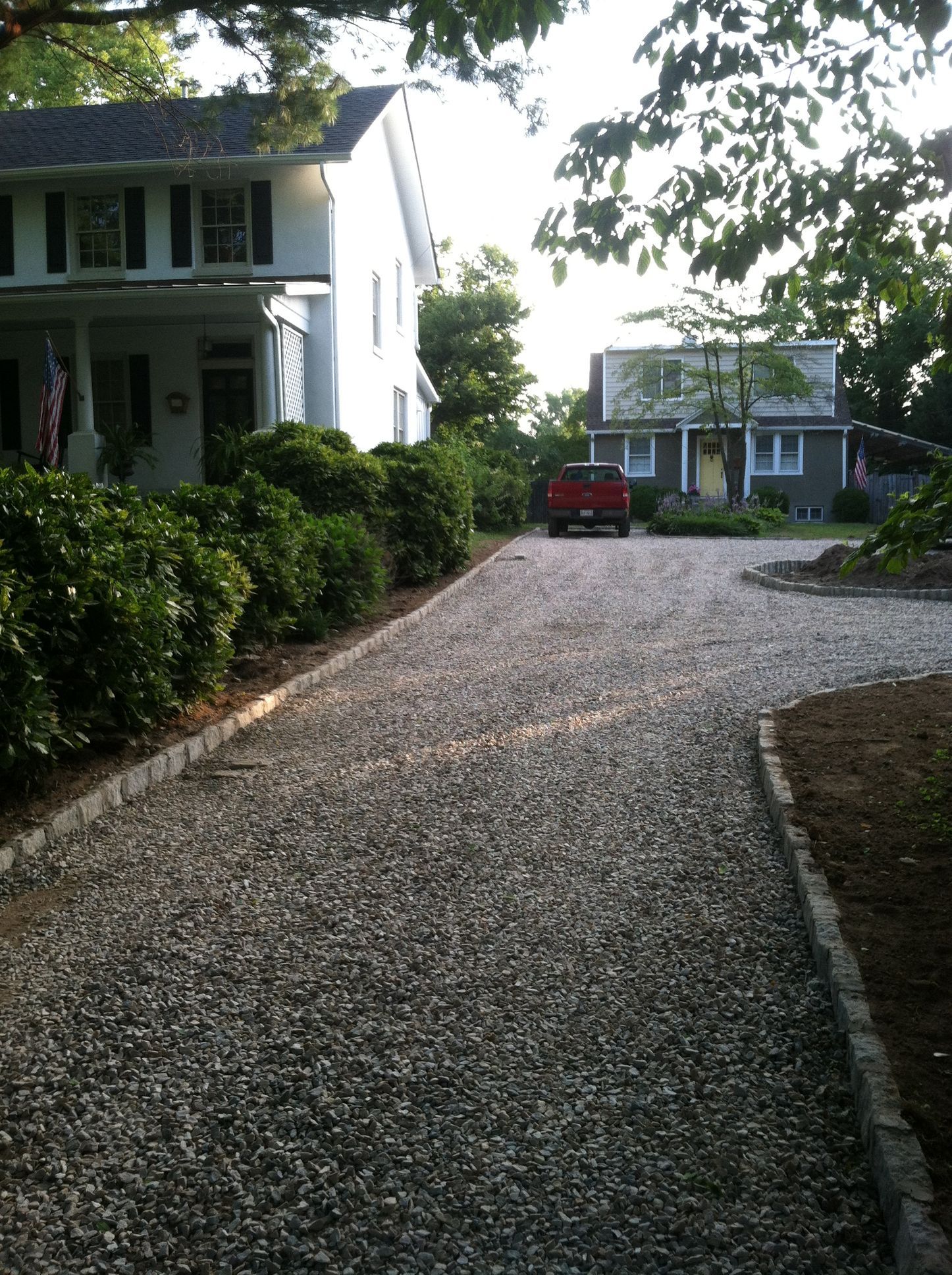 Gravel driveway beside a white house, leading to a small red car and another house in the distance