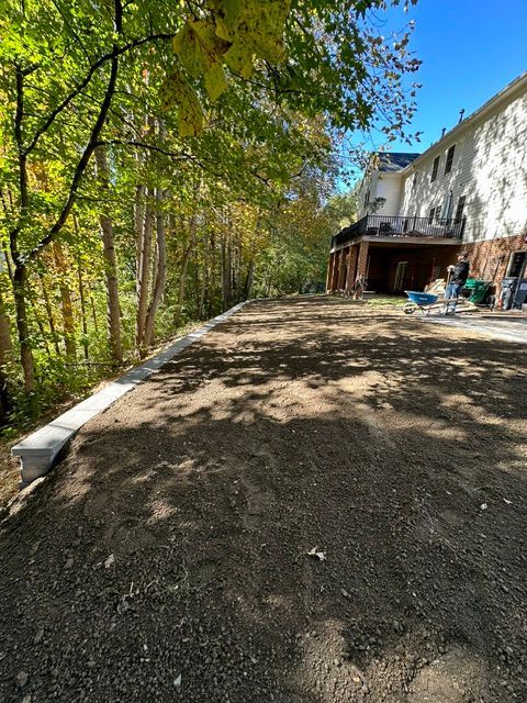 Gravel driveway beside a wooded slope and a white house in bright sunlight