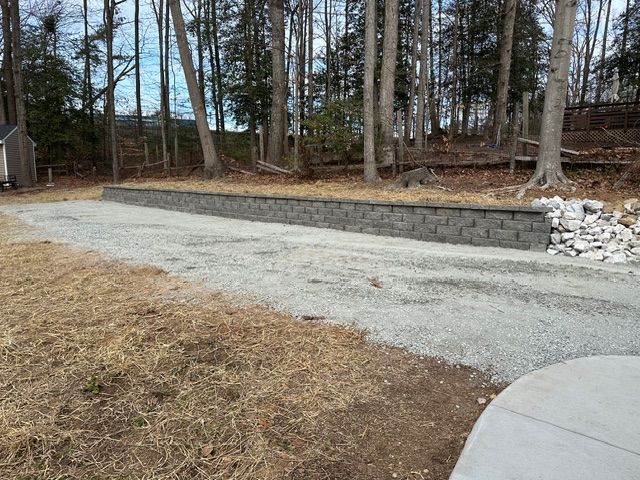 Gravel driveway beside a stone retaining wall and leafless trees in a wooded yard