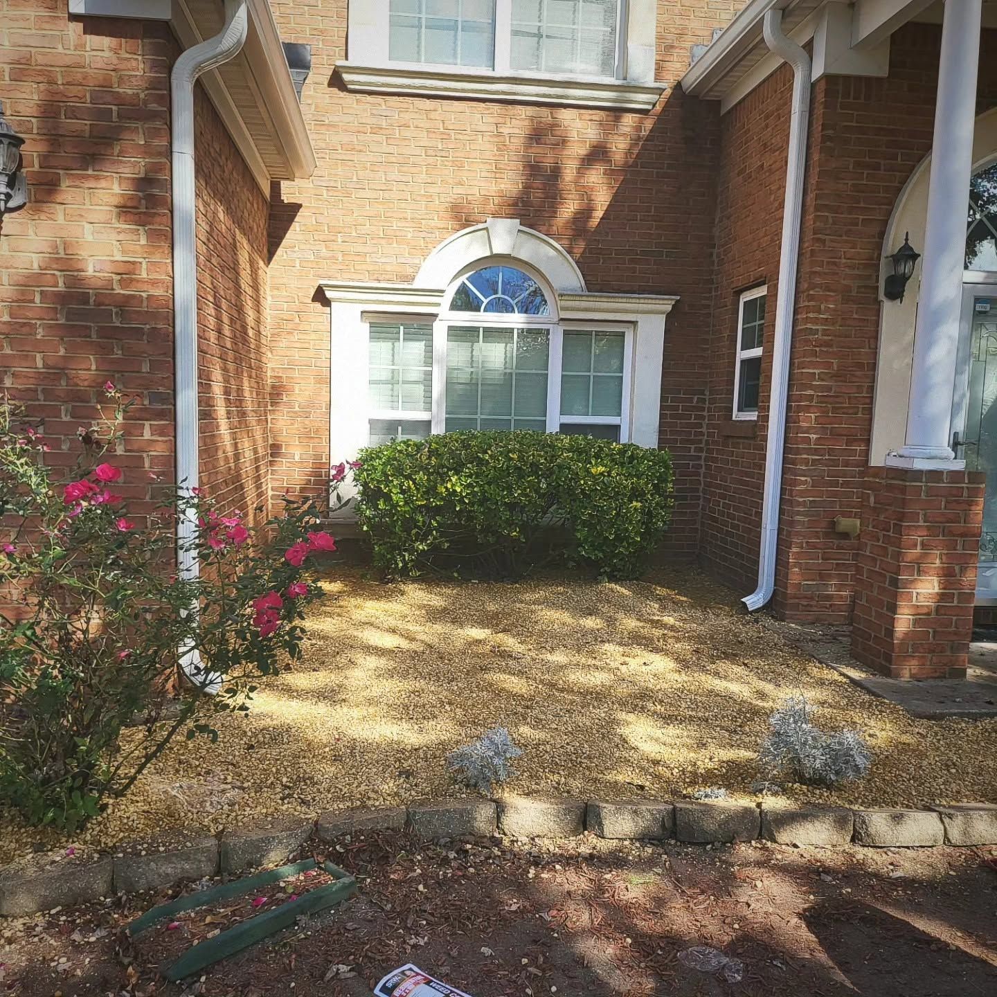 Brick townhouse front entry with white-trimmed window, gravel path, shrubs, and a red flowering bush.