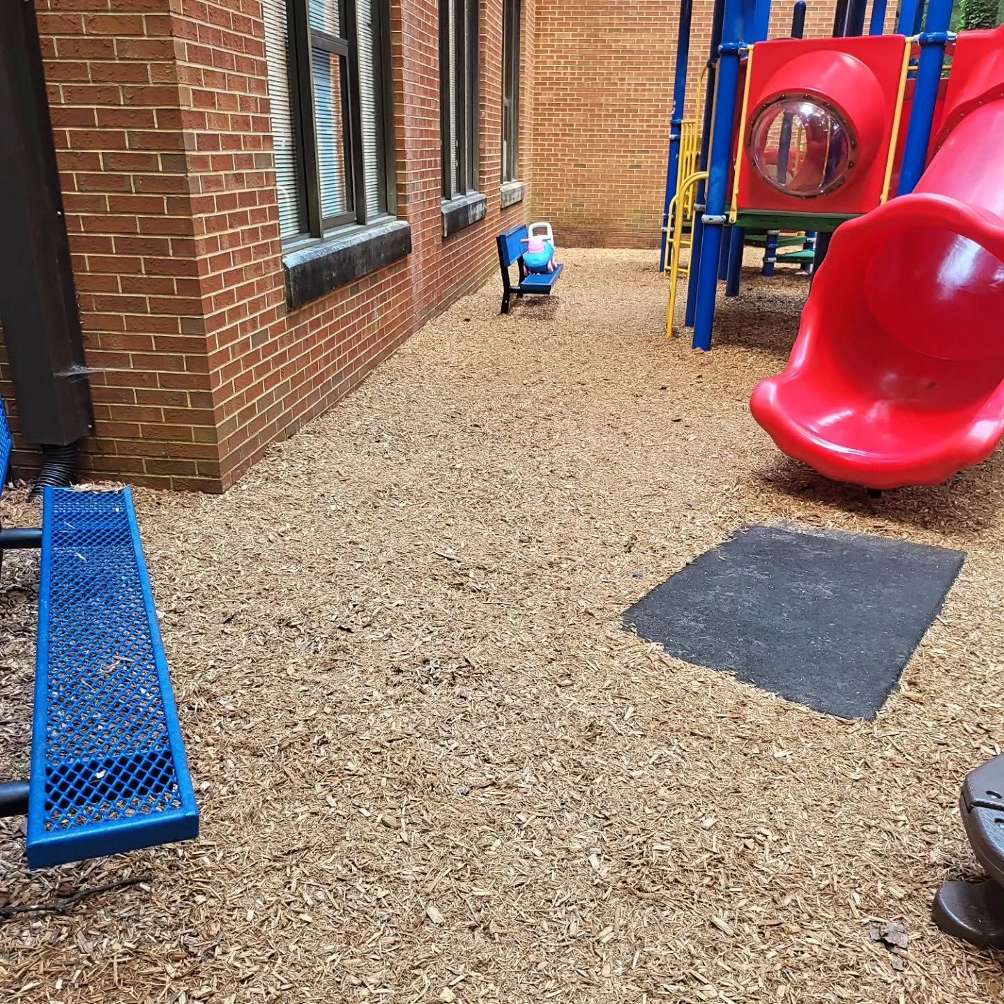 Outdoor playground beside a brick building with red slide, blue climbing frame, and gravel ground.
