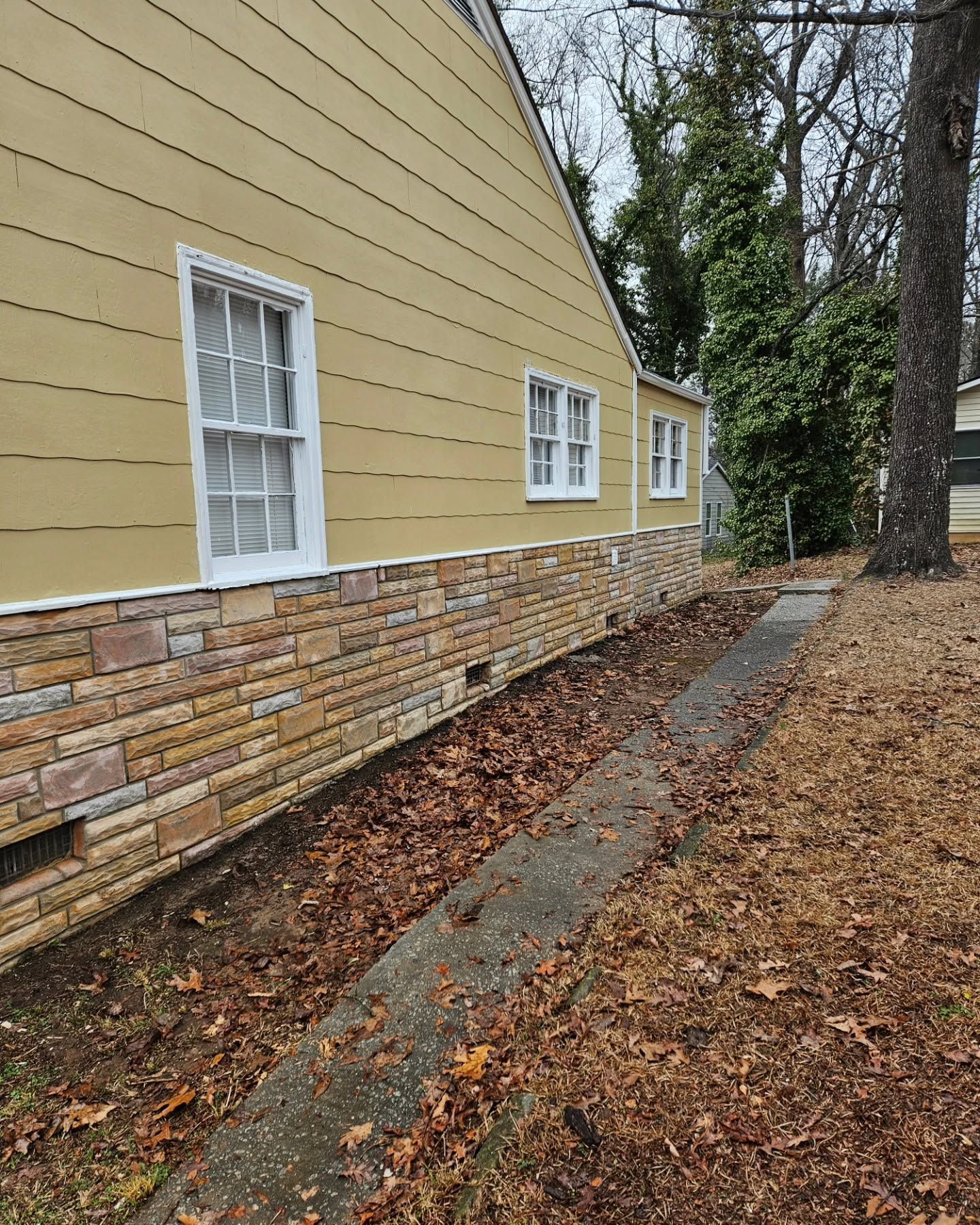 Side of yellow house with white windows and stone base beside a leaf-covered walkway and trees