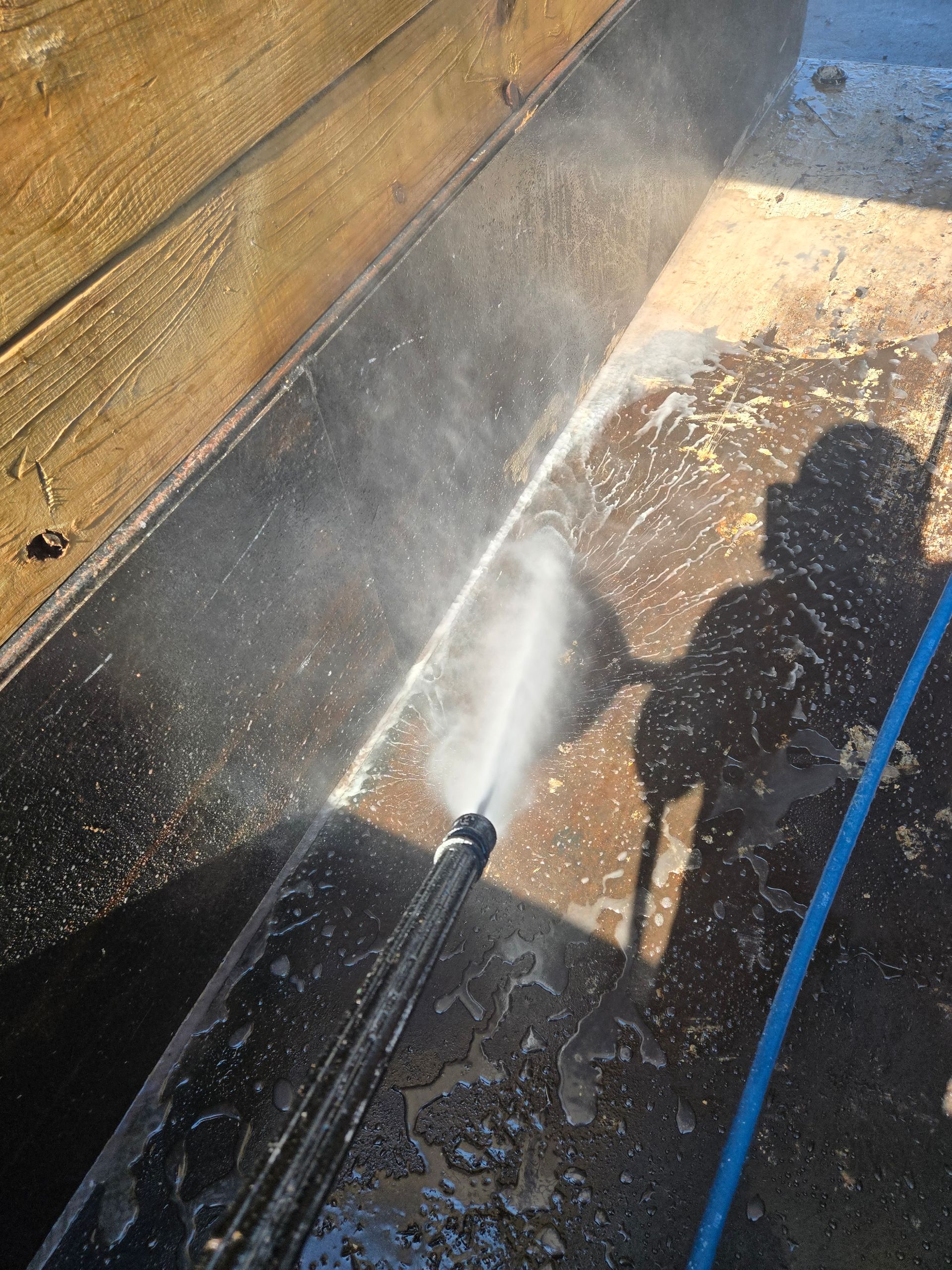 Pressure washing a sunlit wooden deck, with water spraying across the surface and a blue hose visible