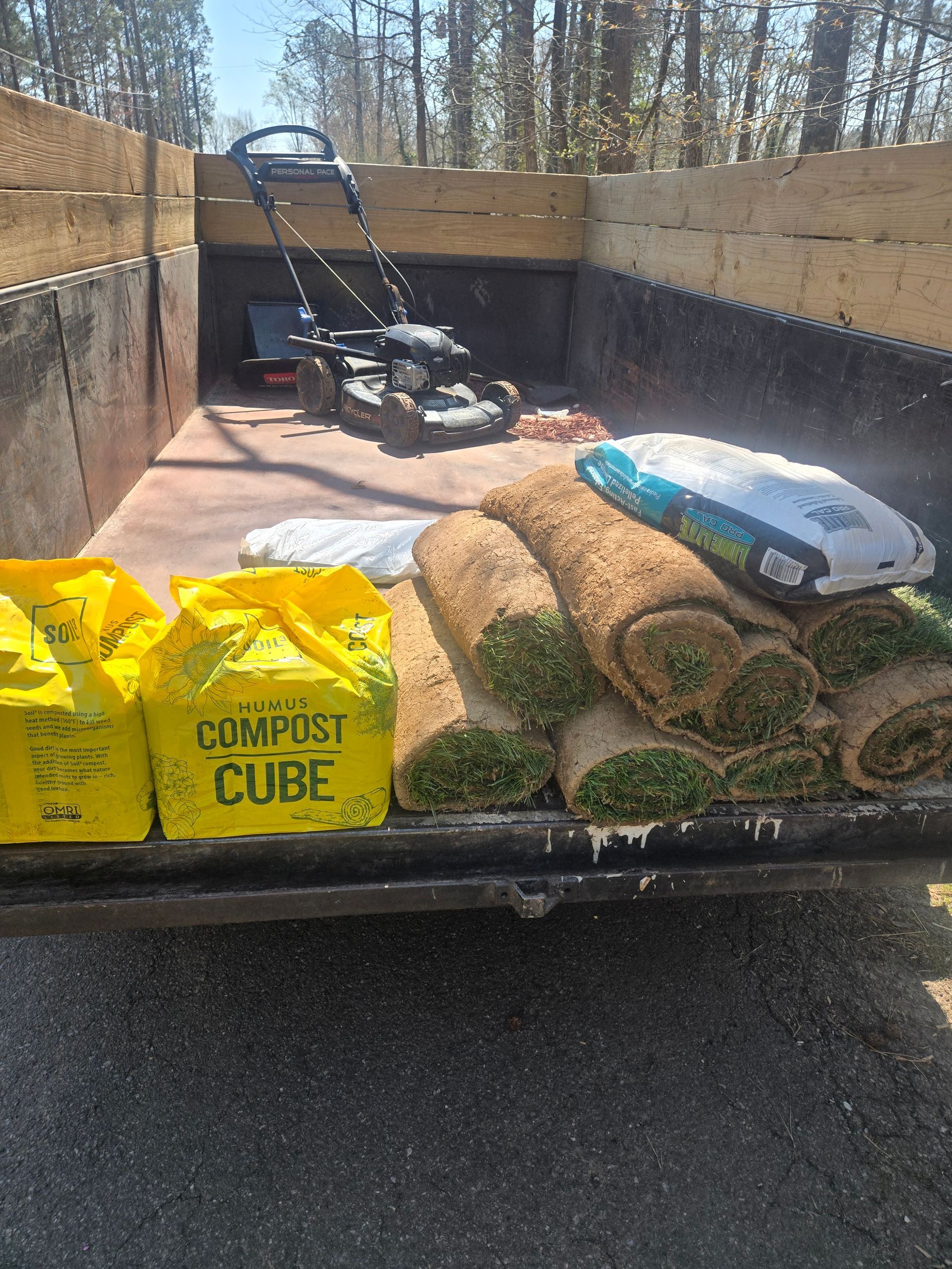 Landscape trailer loaded with lawn care supplies, mulch bags, and a push mower in a sunny driveway.