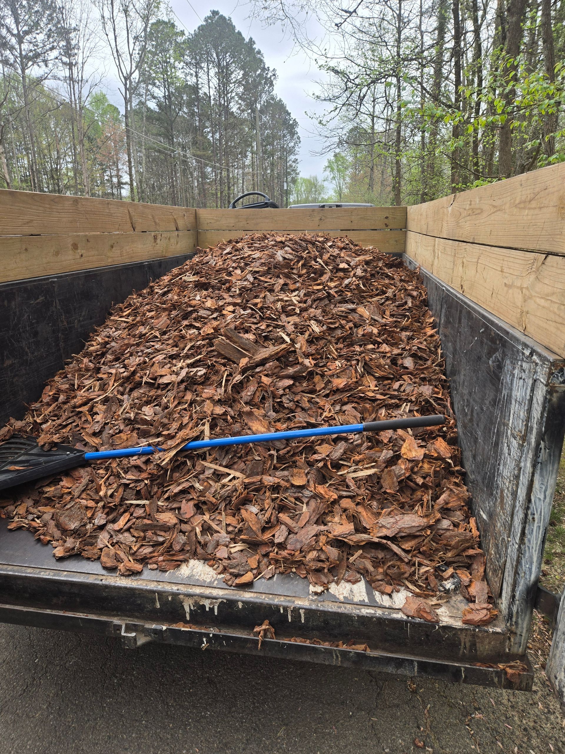 Leaves piled in a trailer bed with a blue rake handle, outdoors among trees.