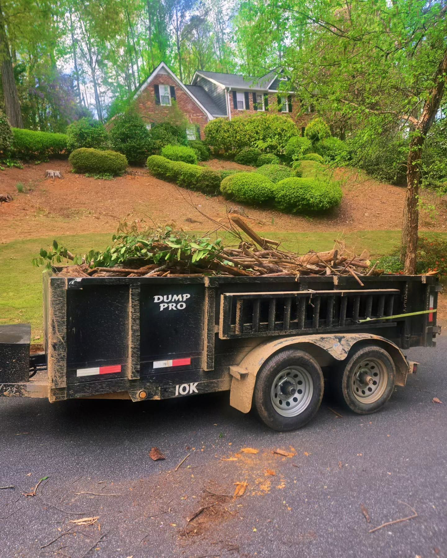 Dump trailer with yard debris parked by a house in a wooded neighborhood.