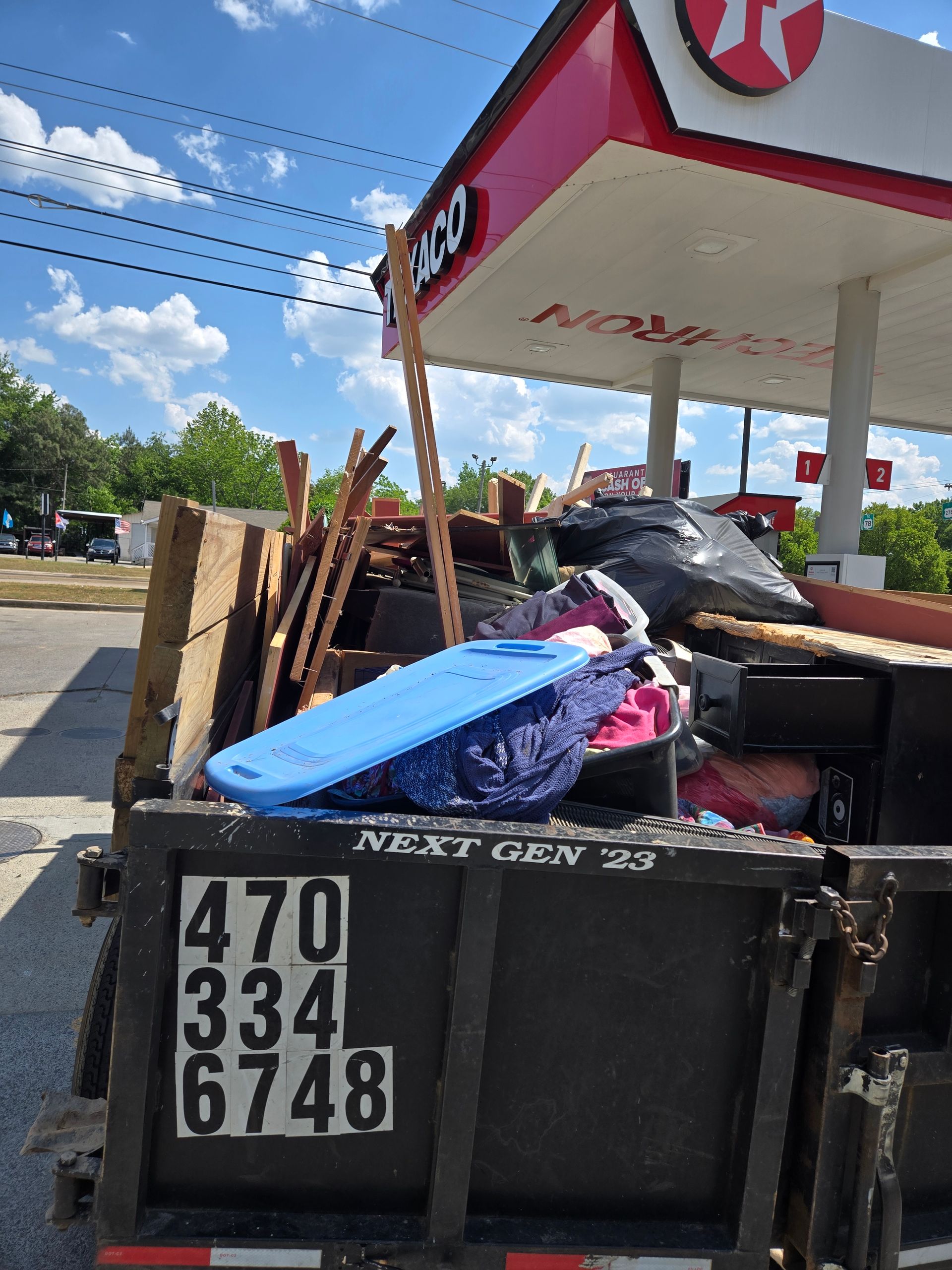 Overflowing trash bin with wood, bags, and a blue cooler at a gas station curb