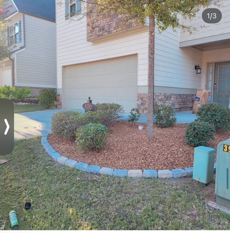 Front yard landscaping with shrubs and mulch beside a house, bordered by blue stones near a garage.