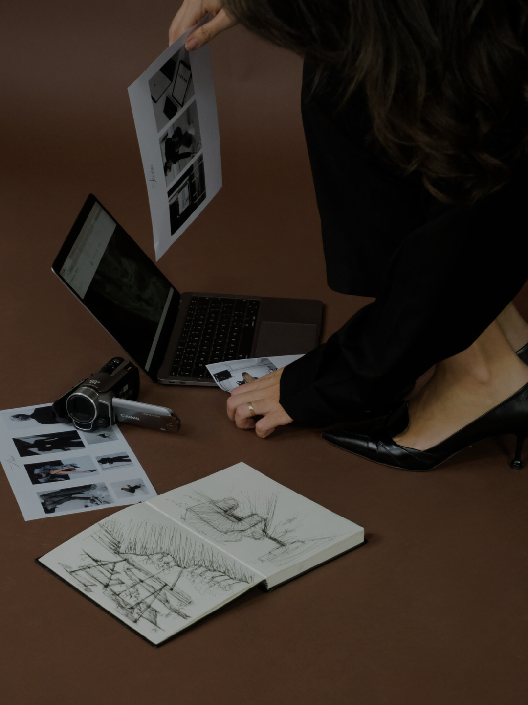 Een persoon die fotoafdrukken naast een laptop en een schetsboek op een houten vloer rangschikt.