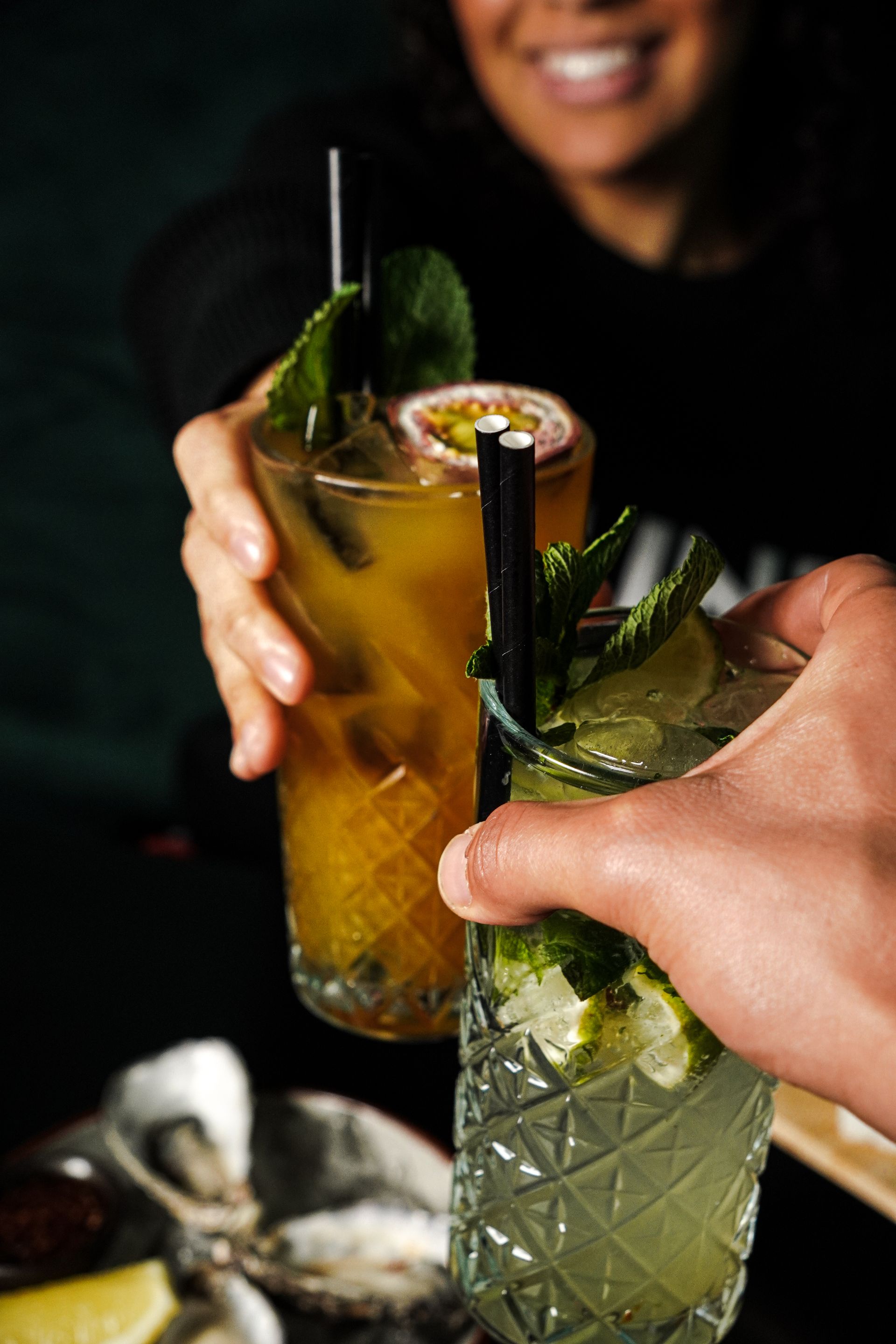 Twee cocktails, een amberkleurige en een groene, klonken tegen elkaar, met een glimlachend persoon wazig op de achtergrond.