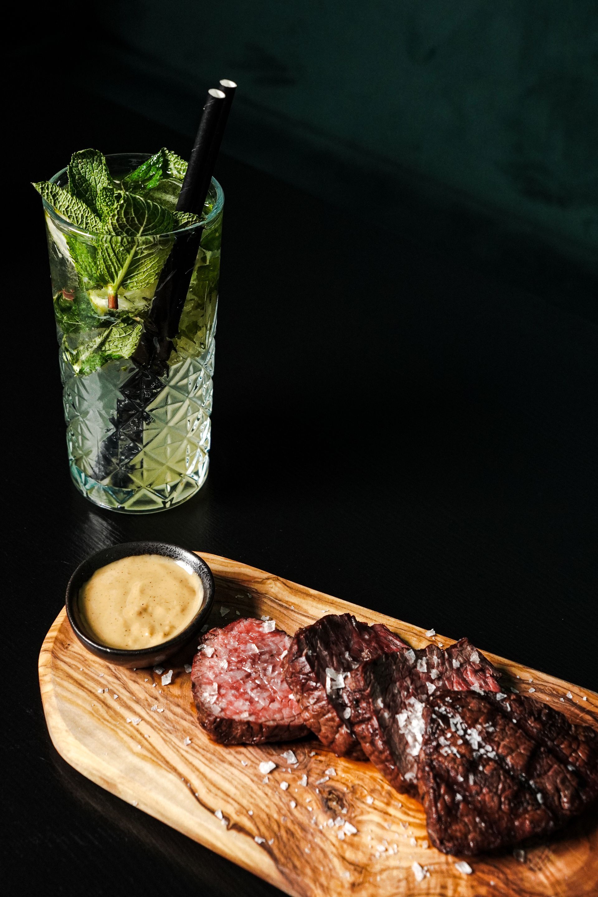 Biefstuk op een houten plank met saus, naast een muntcocktail in een hoog glas.