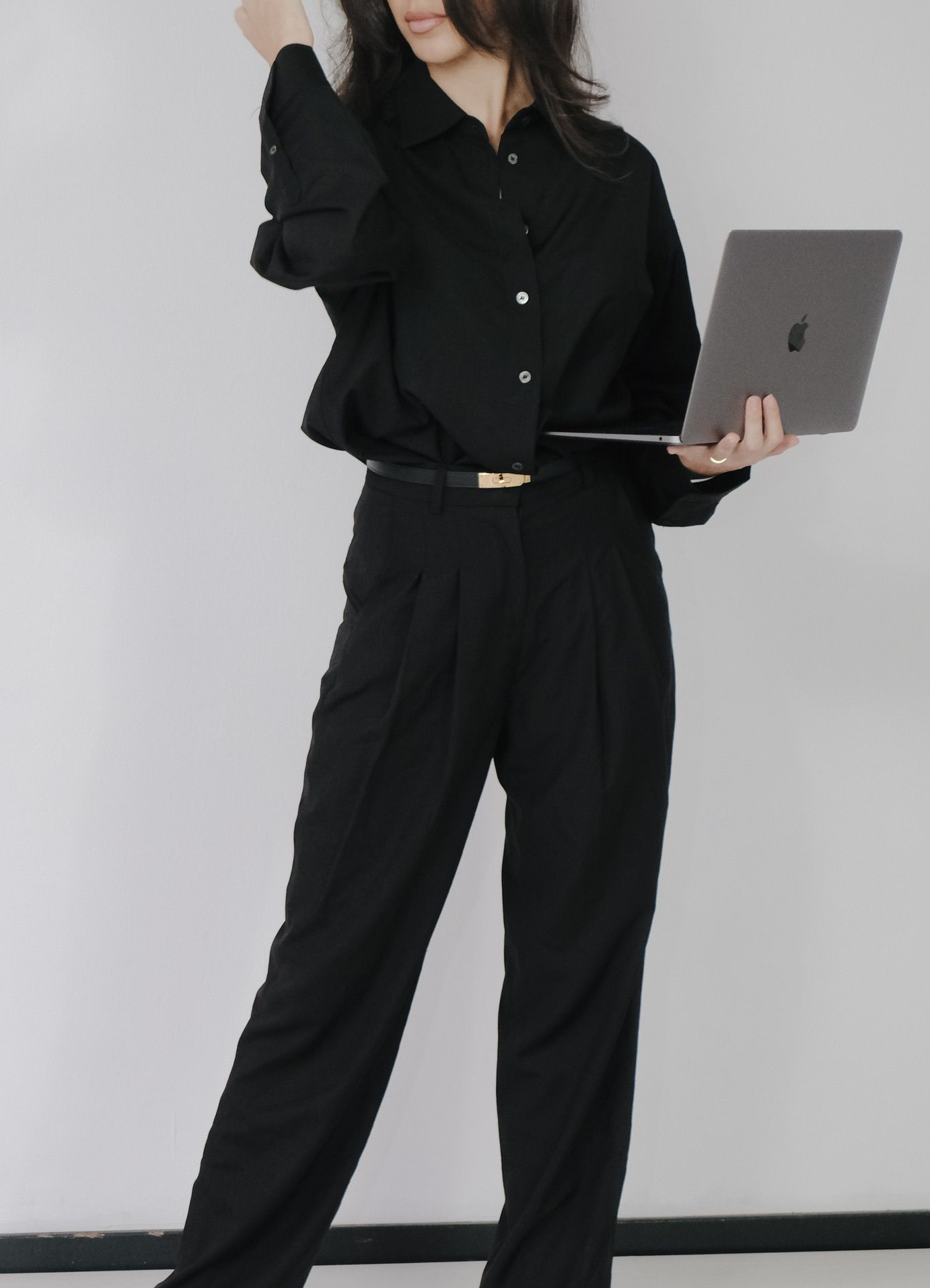 Persoon in zwarte kleding met een opengeklapte laptop in de hand, staand tegen effen grijze achtergrond.