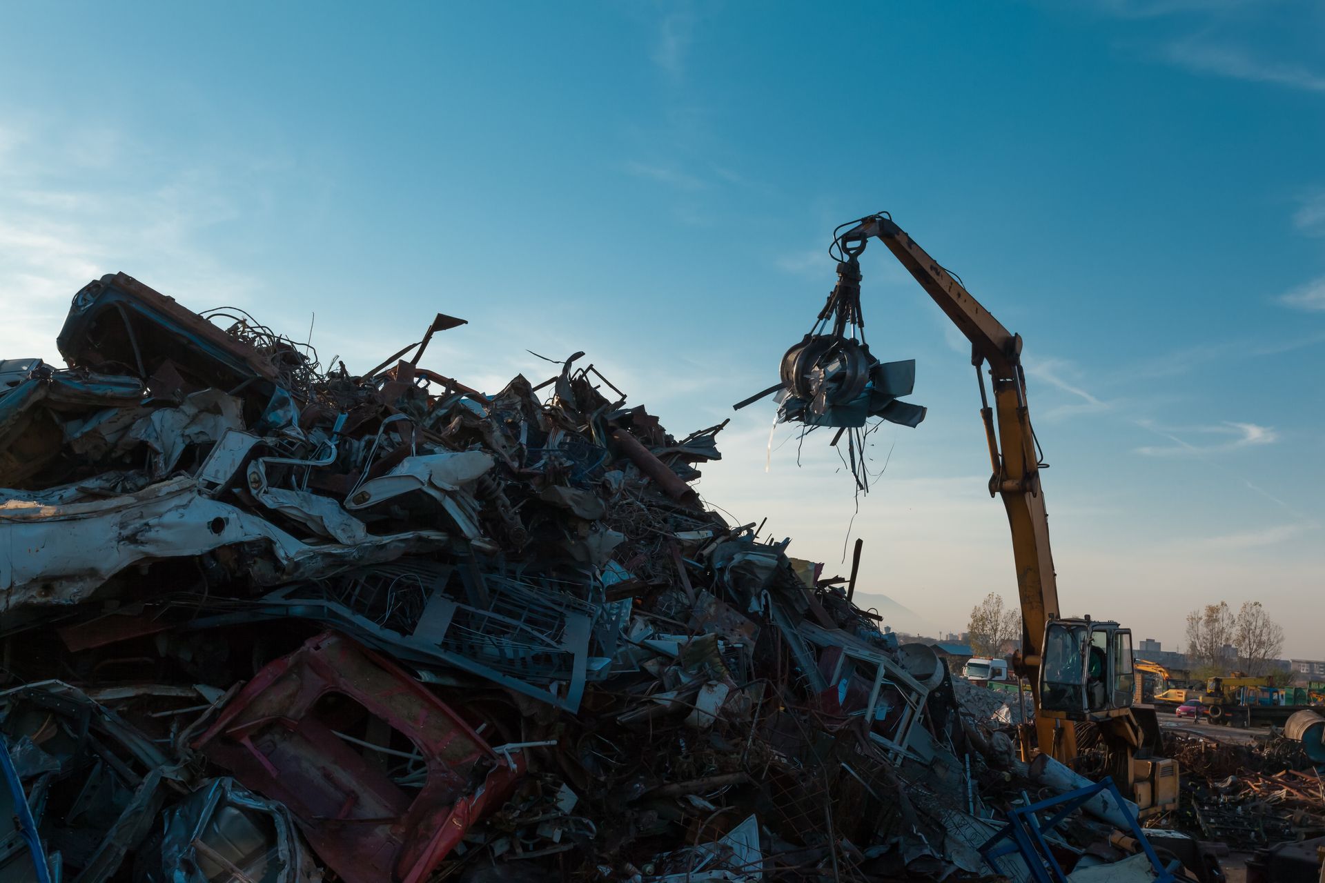 Crane grapple lifts tangled scrap from a towering pile of crushed cars and twisted metal at sunset.