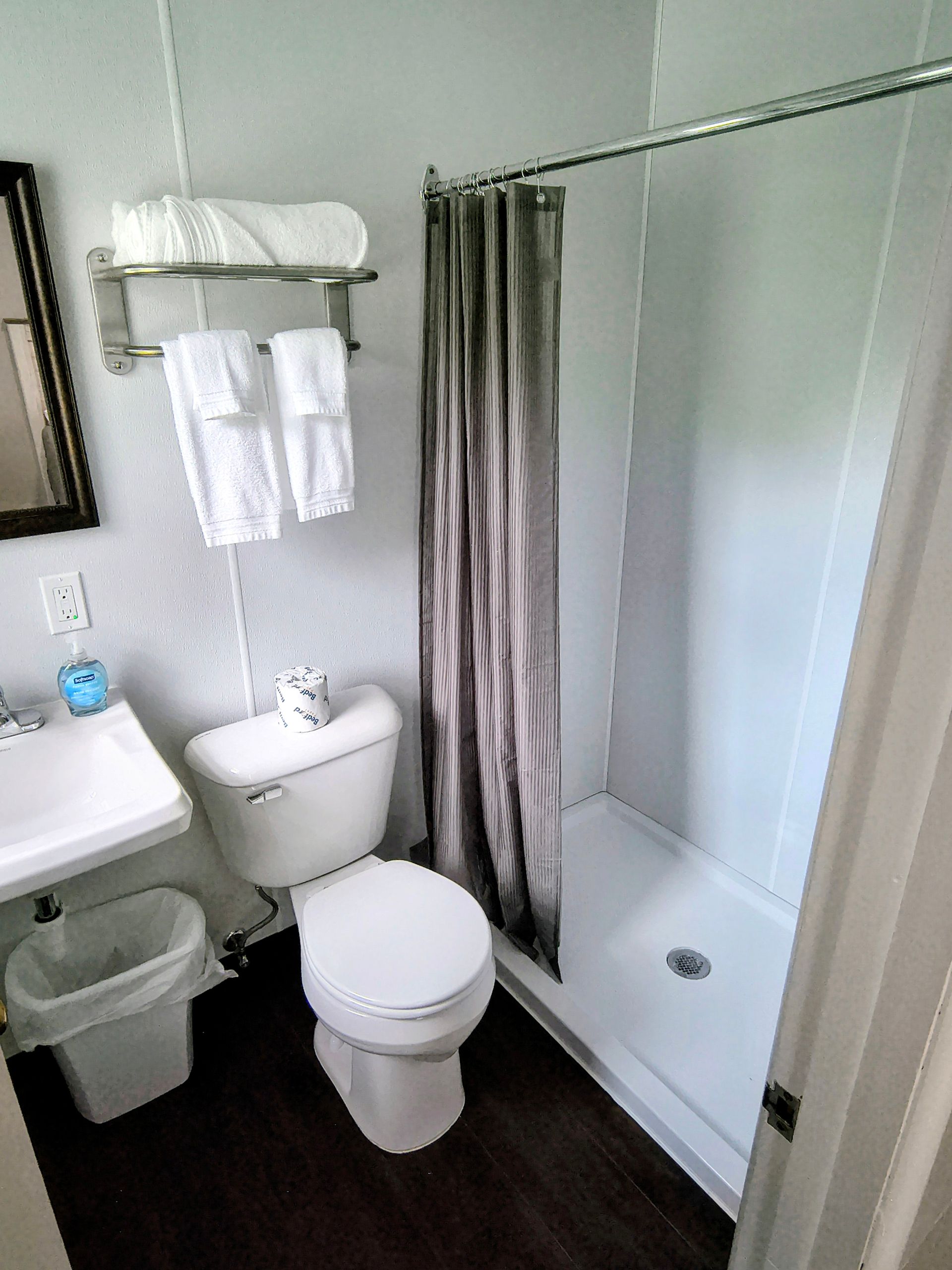 Small motel bathroom with toilet, sink, shower, and towels; dark wood-look floor, light walls.