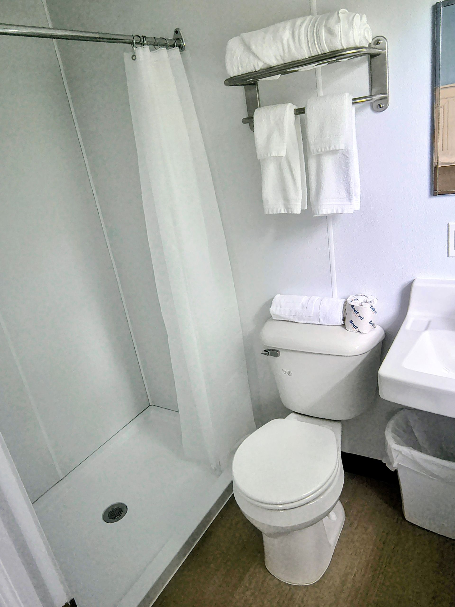 Small, clean bathroom with shower, toilet, sink, and towels. White walls and a light-colored floor.