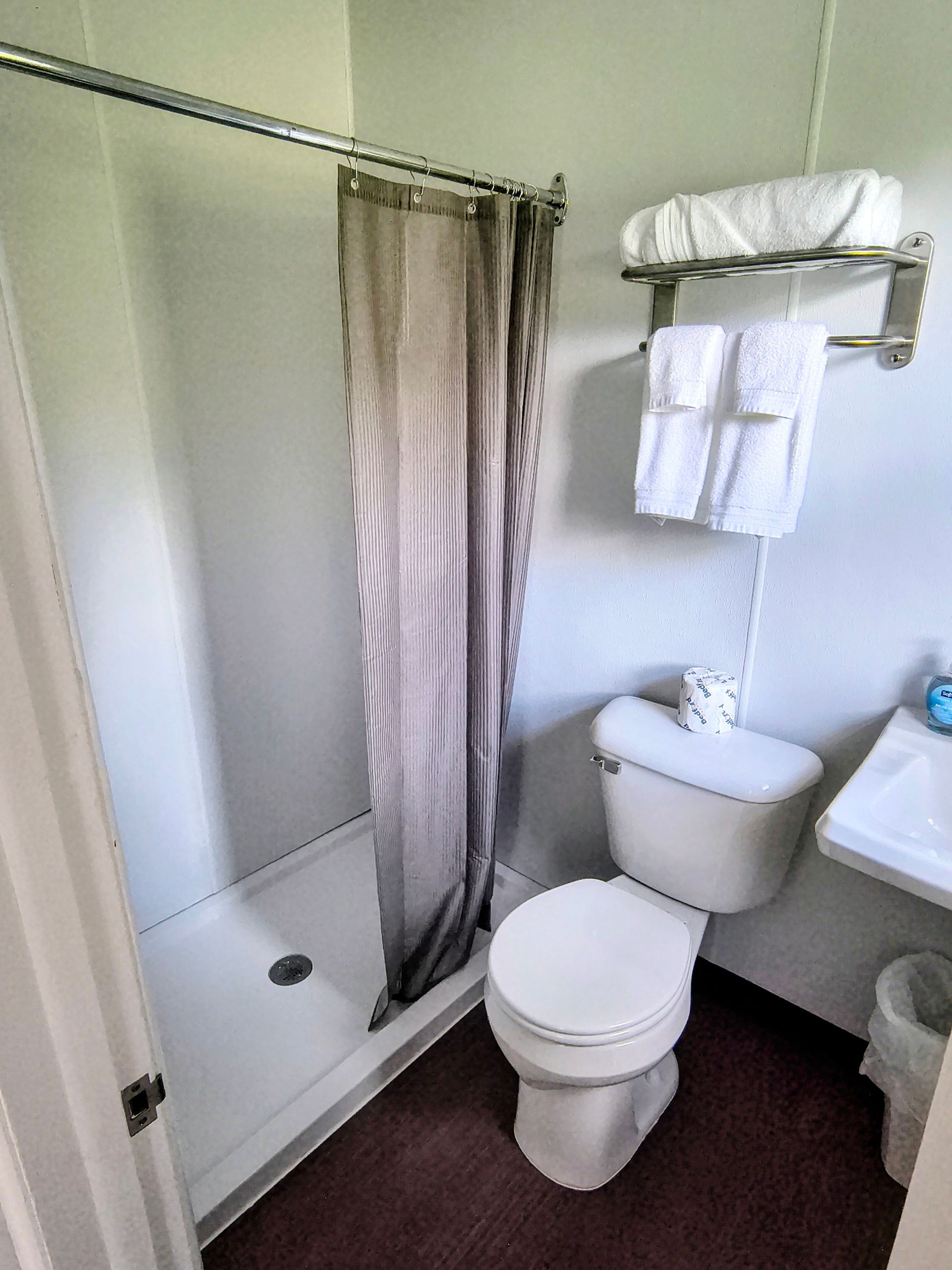 Small motel bathroom with shower, toilet, sink, and towels. White fixtures, red floor, neutral color shower curtain.