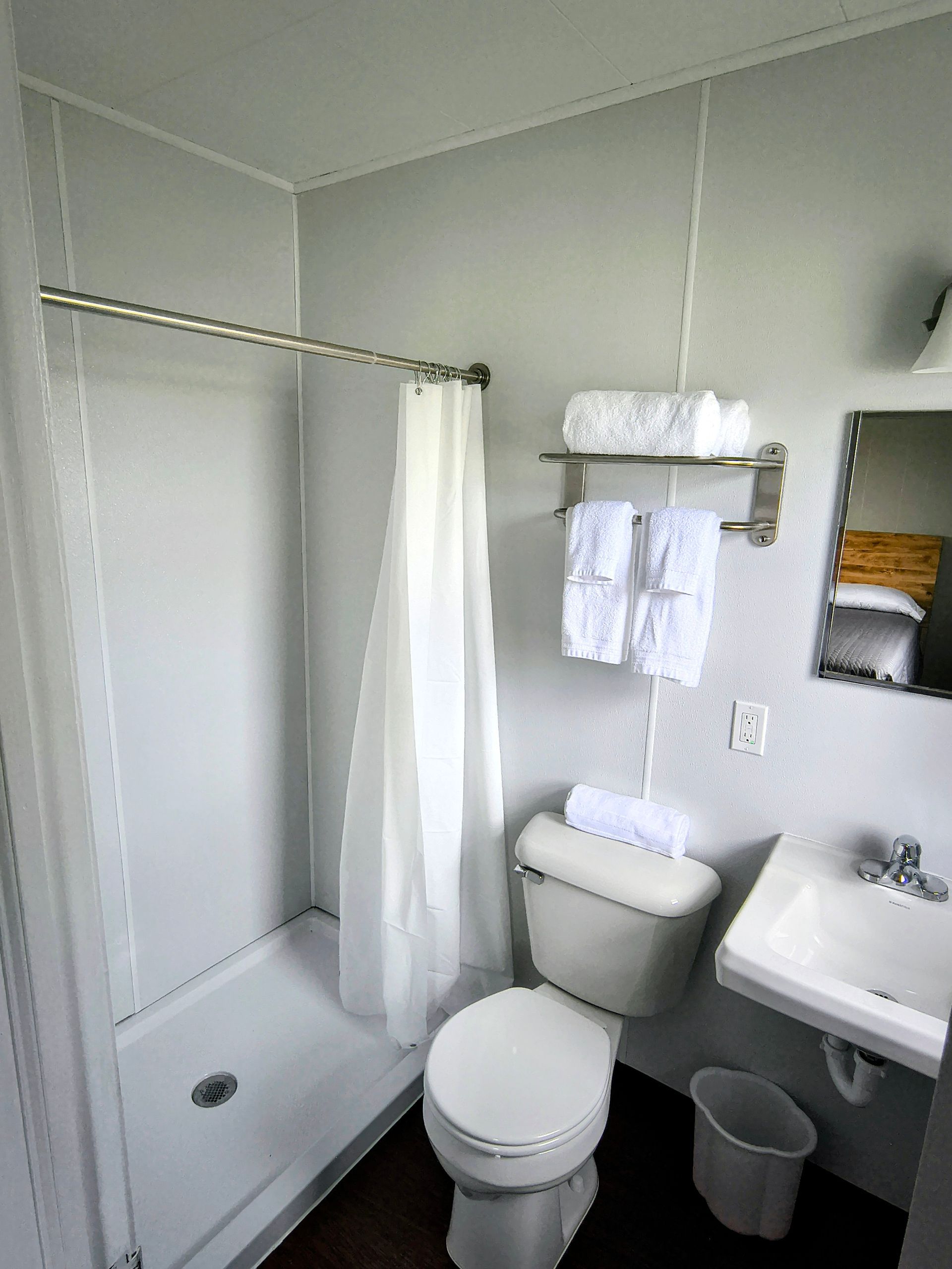Small, plain bathroom with a shower, toilet, sink, and towels. White walls and fixtures, dark floor.