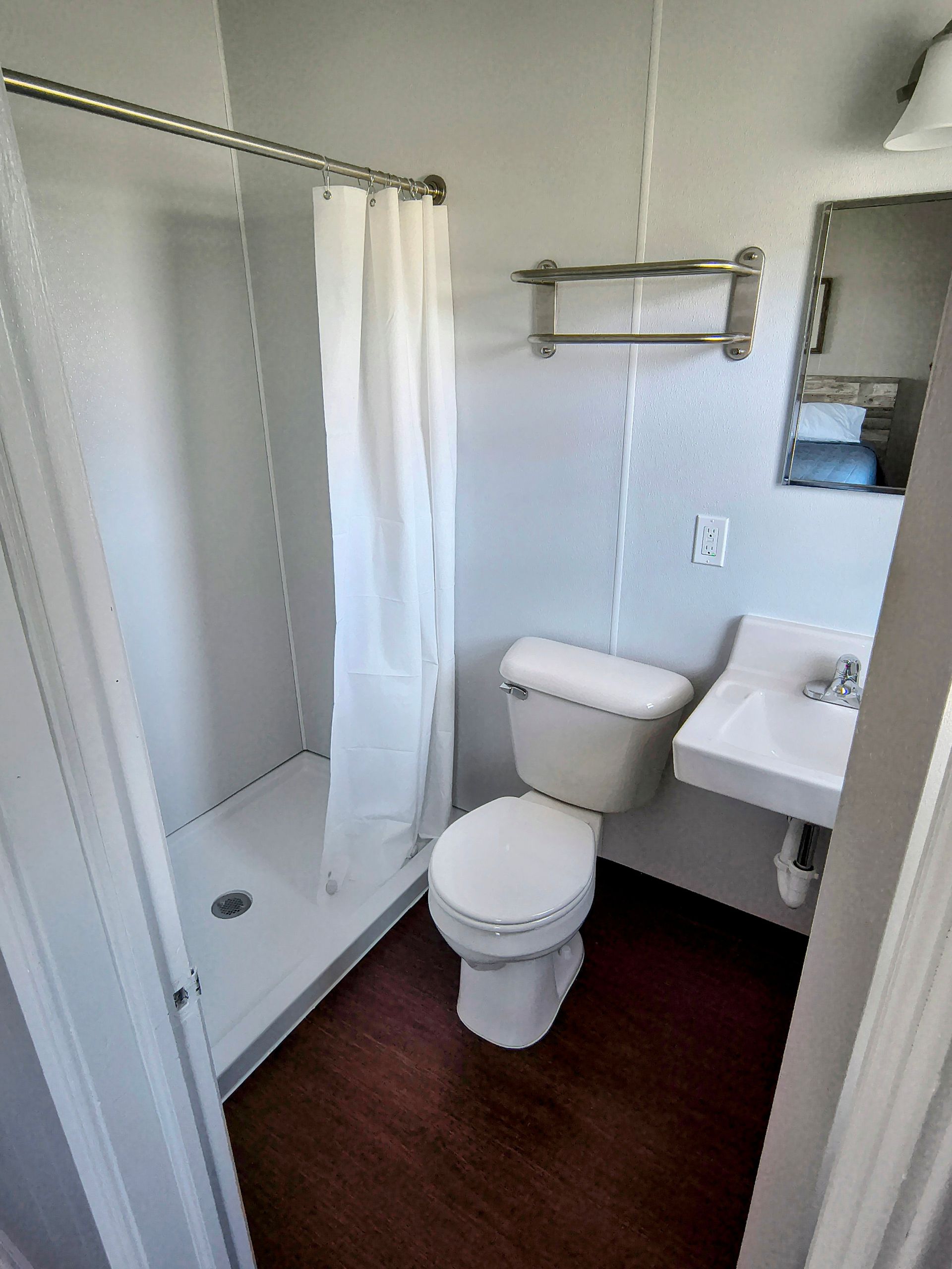 A small, white-walled bathroom with a shower, toilet, and sink. The floor is dark wood-toned.